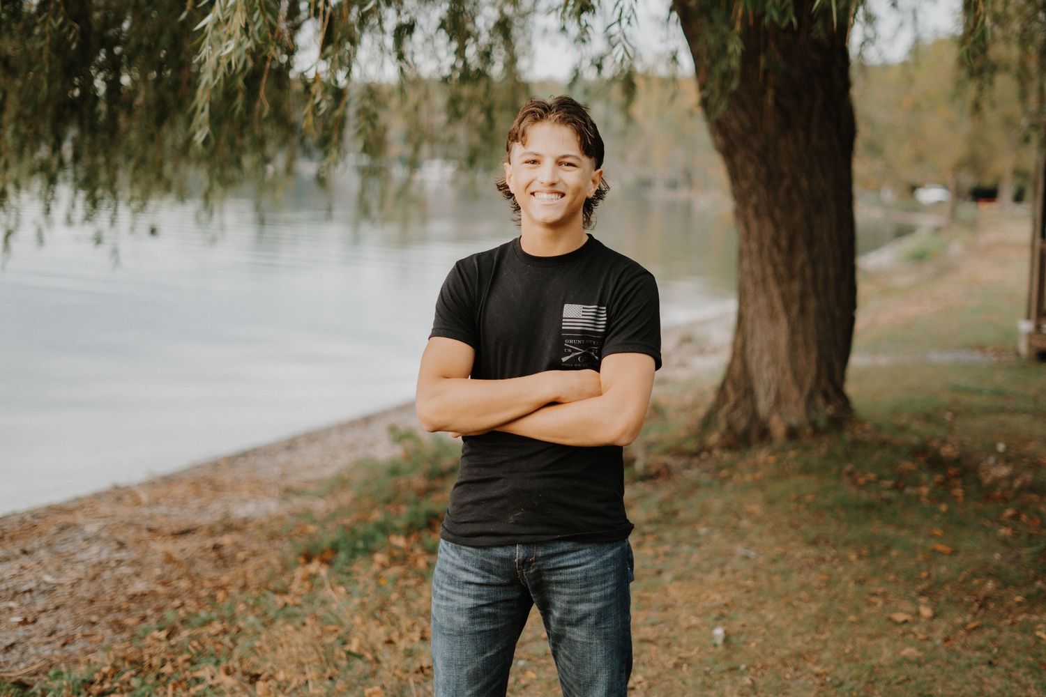 An outdoor senior portrait of a boy standing with crossed arms near a lake and willow trees.