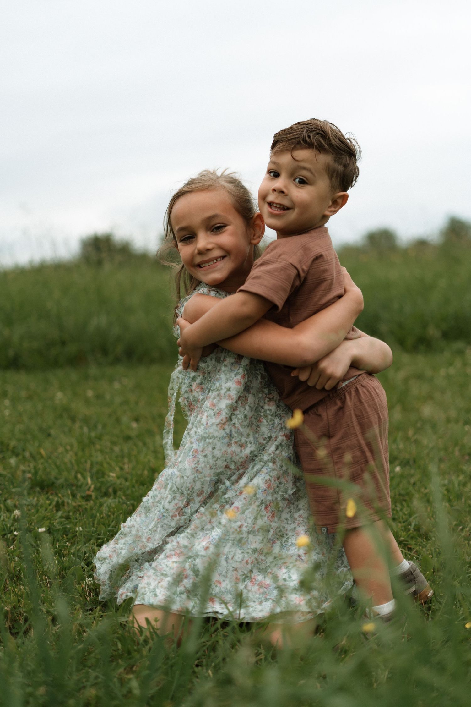 Two children embrace and play together in a grassy summer field, with one wearing a floral dress.
