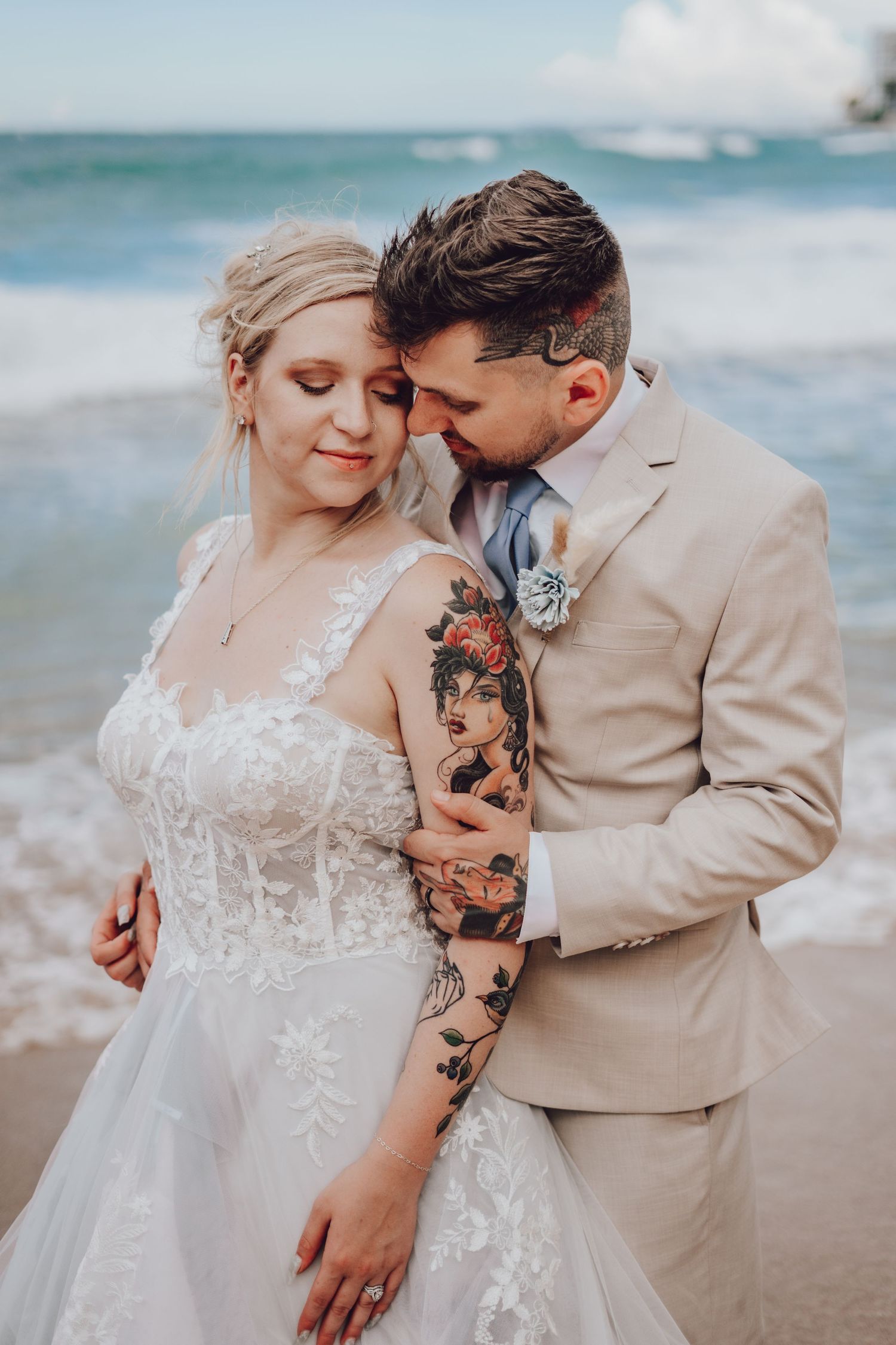 A couple embraces on a beach, with the groom in tan suit showing tattoo sleeve.