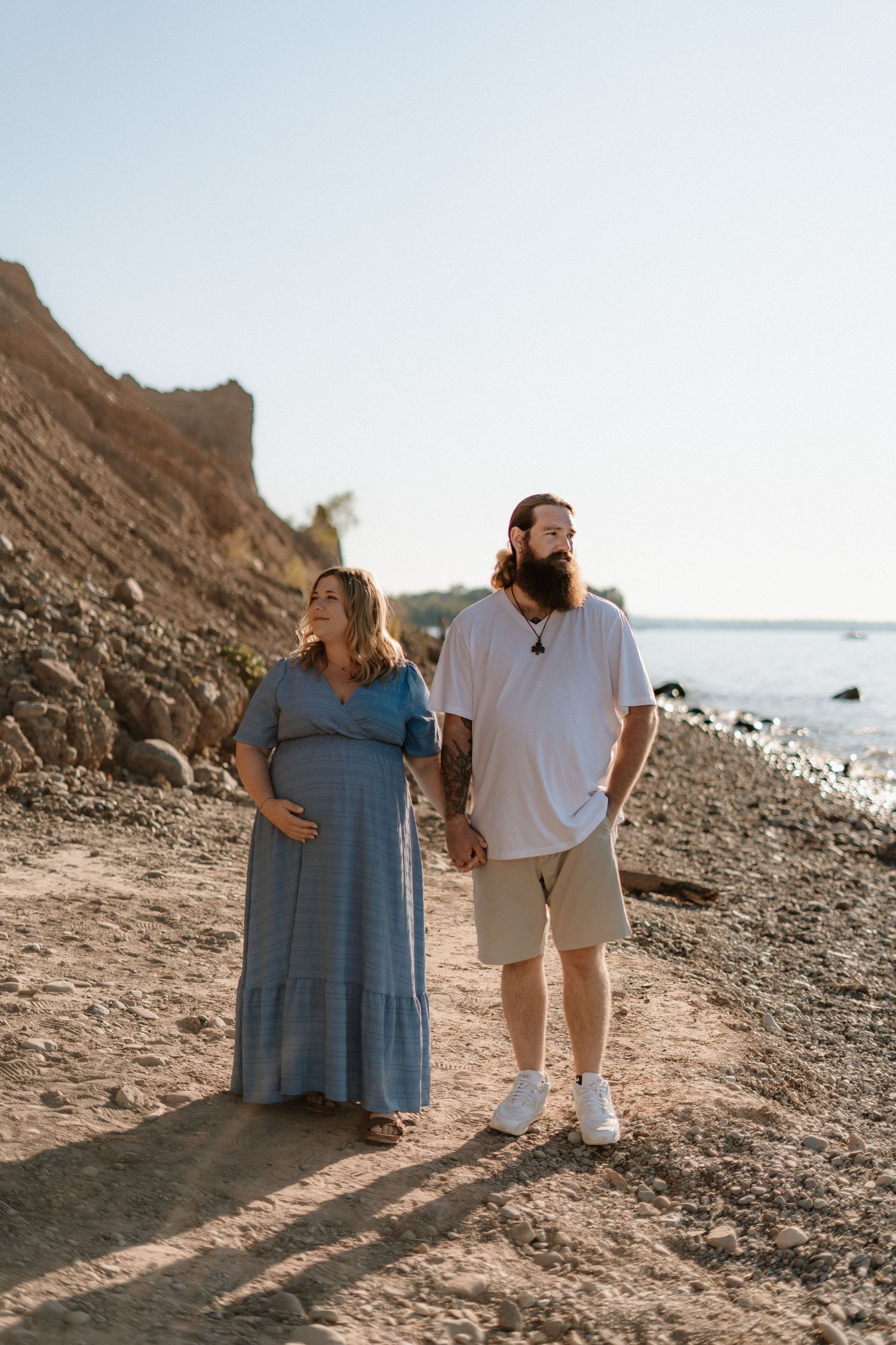 A couple stands together on a rocky beach at sunset casting long shadows on the sand and cliffs.