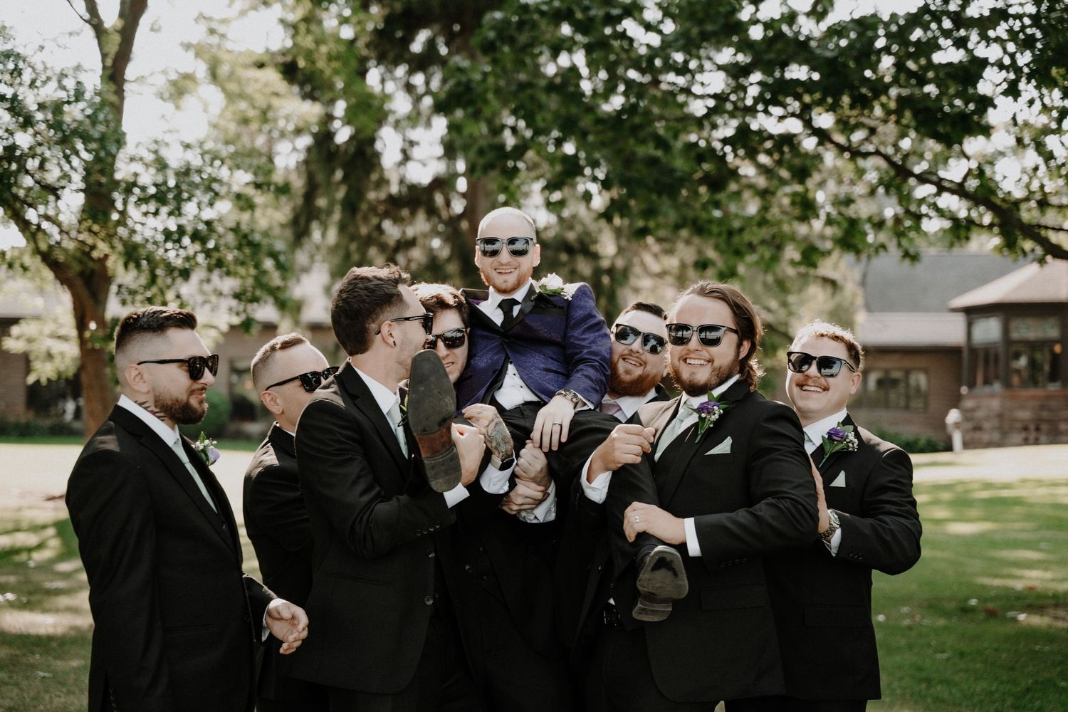 Groomsmen in black suits and sunglasses playfully lift groom during wedding party photos.