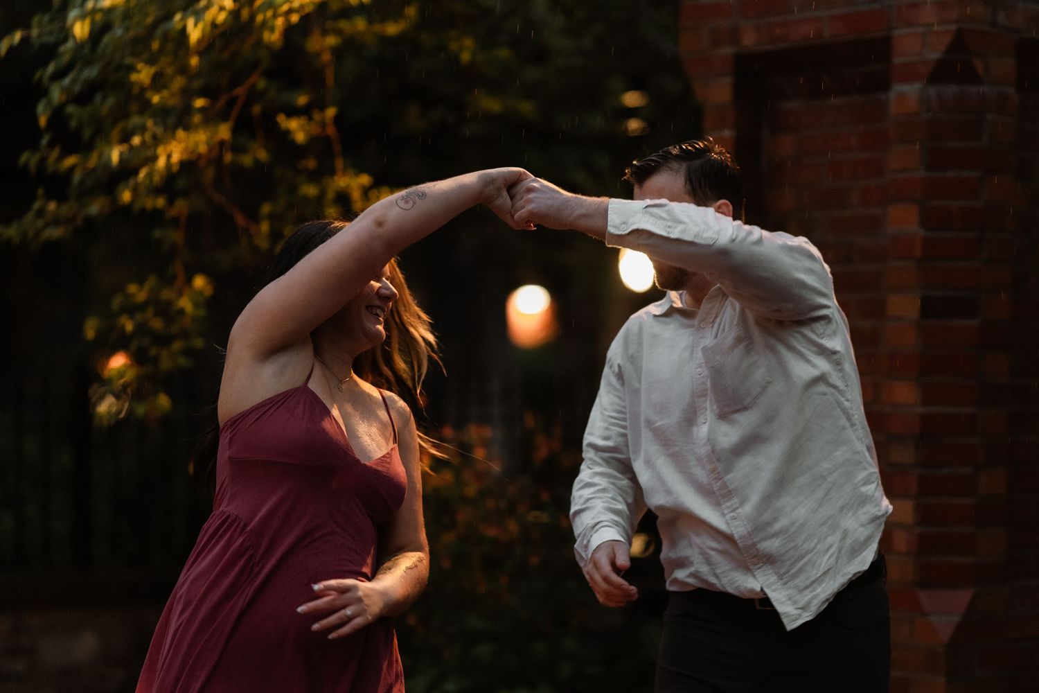 Couple dancing outdoors at dusk, woman in burgundy dress twirling, man in white shirt, warm bokeh lights in background.