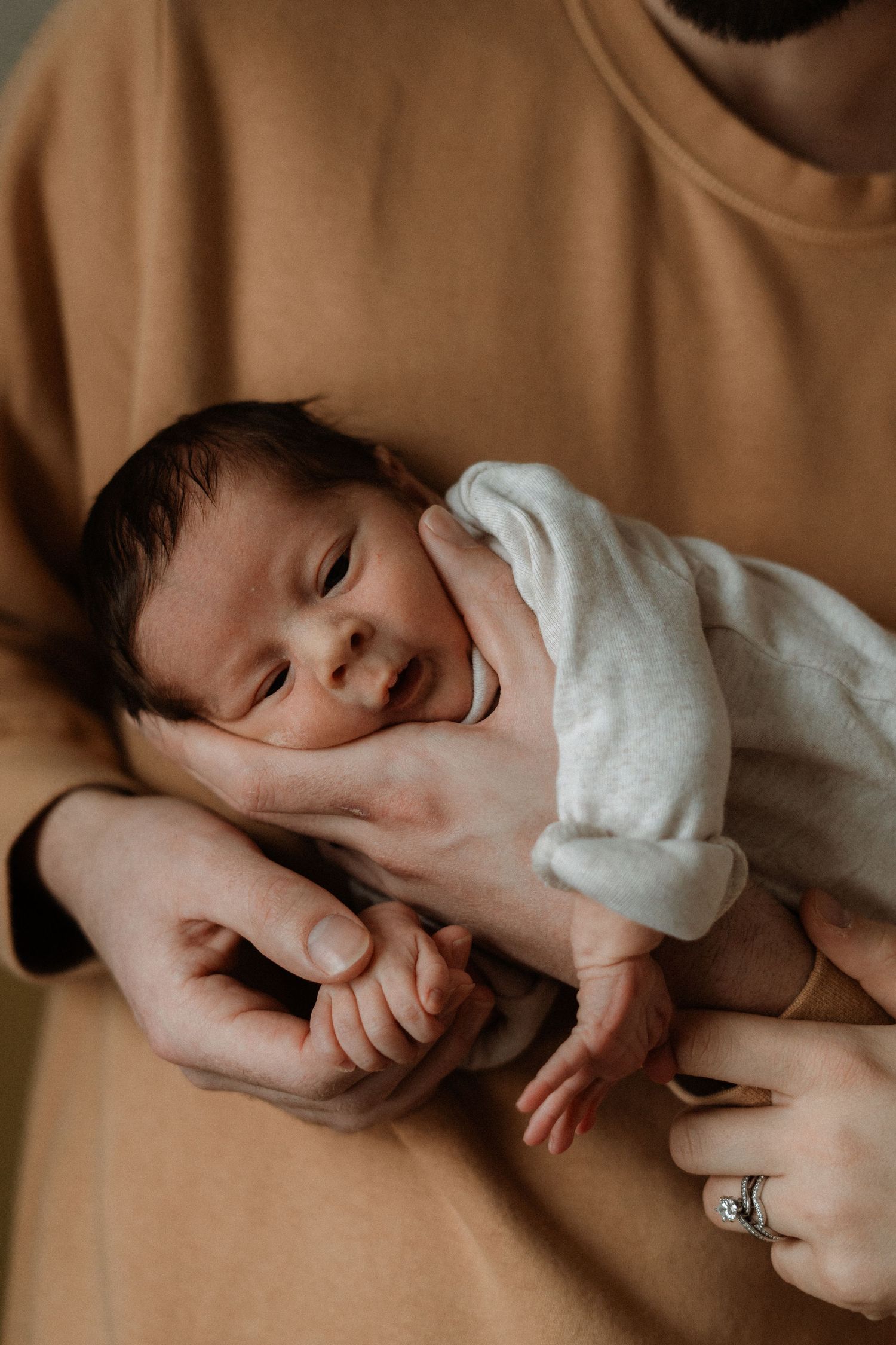 A newborn baby smiling while being cradled in loving arms, wearing a white outfit, held by a parent wearing a gold ring.