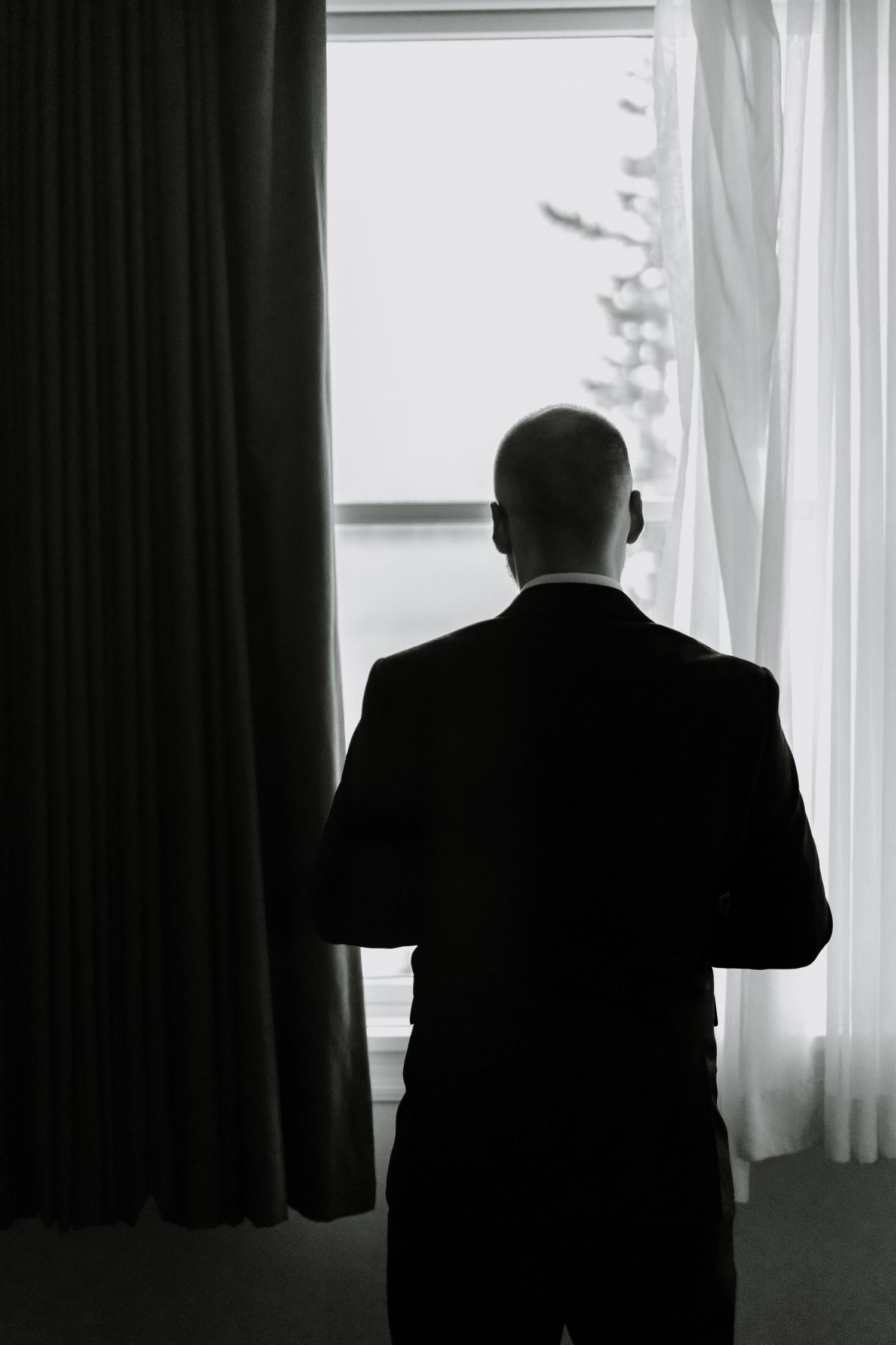 A silhouette of a person in a suit gazing out a window between dark curtains in black and white.