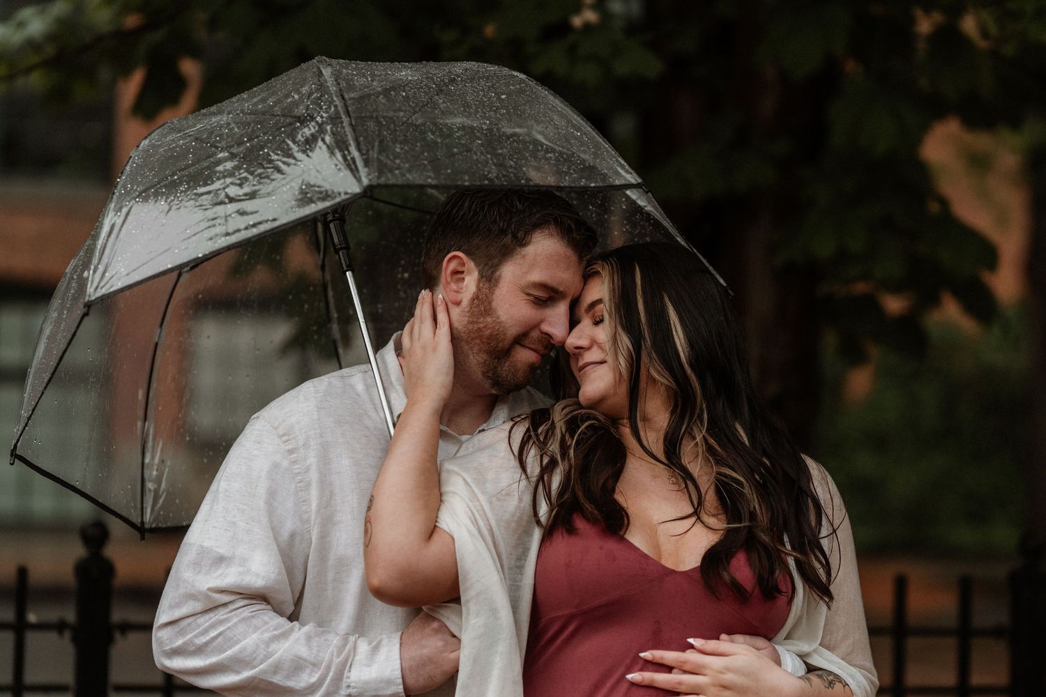 Couple sharing a tender moment under a clear umbrella in the rain, woman in red maternity dress.