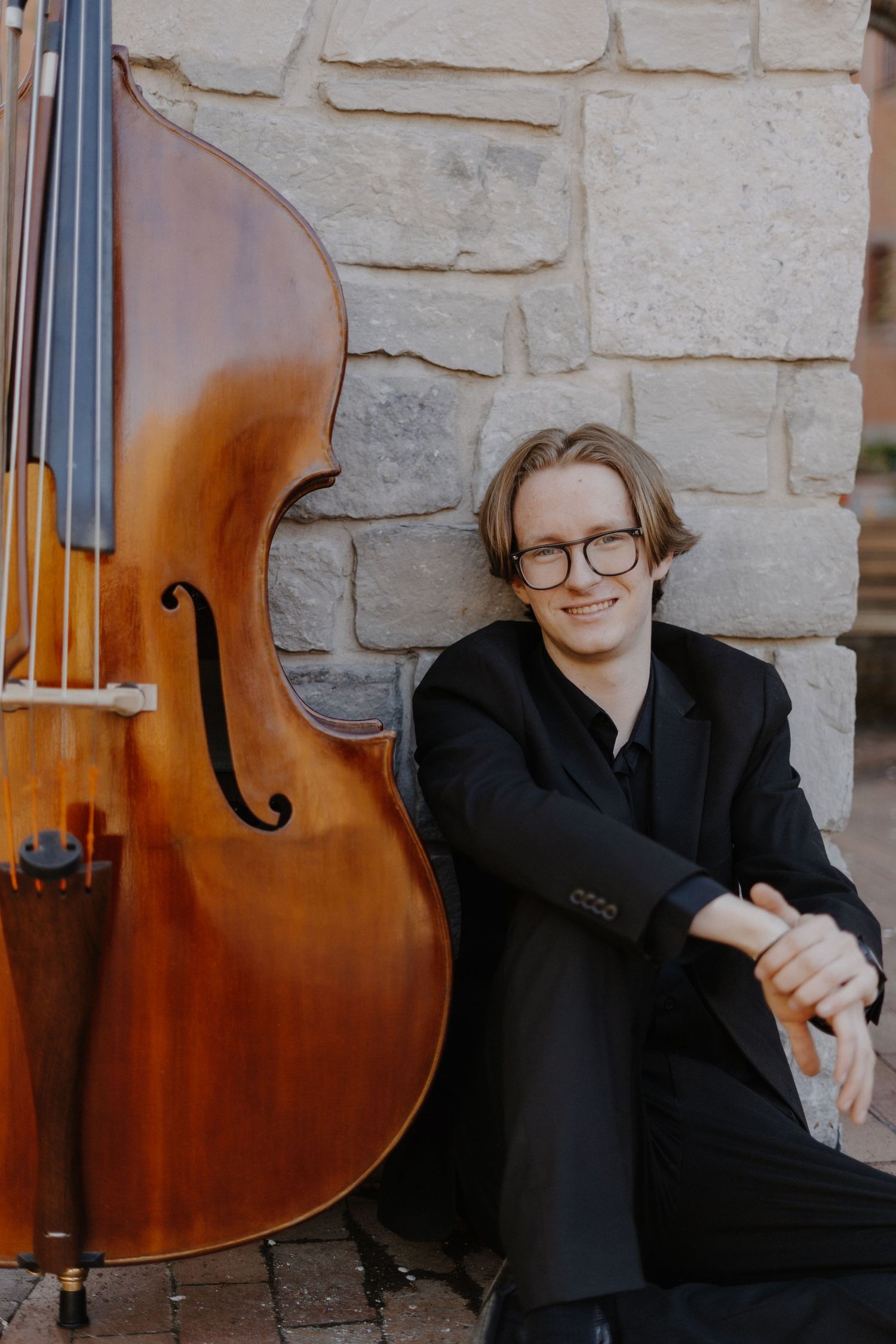 Senior portrait of a boy posing with an upright bass against a stone wall.