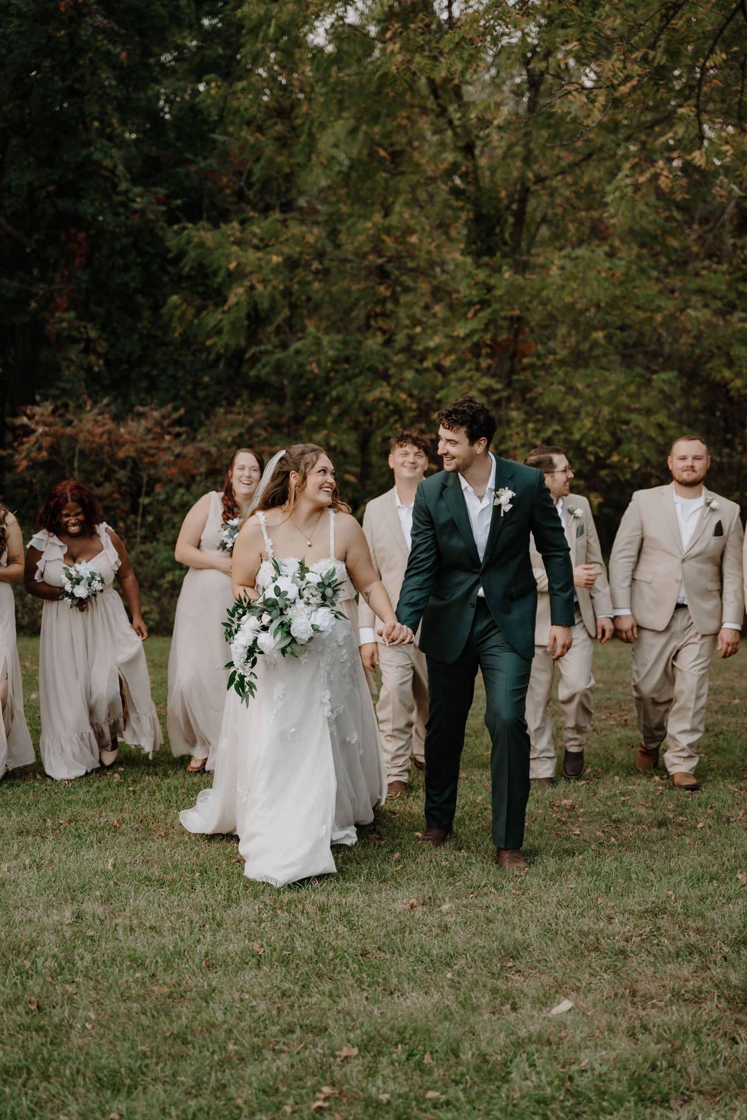 Wedding party walks across grass with bride in white dress and groom in emerald suit accompanied by attendants.