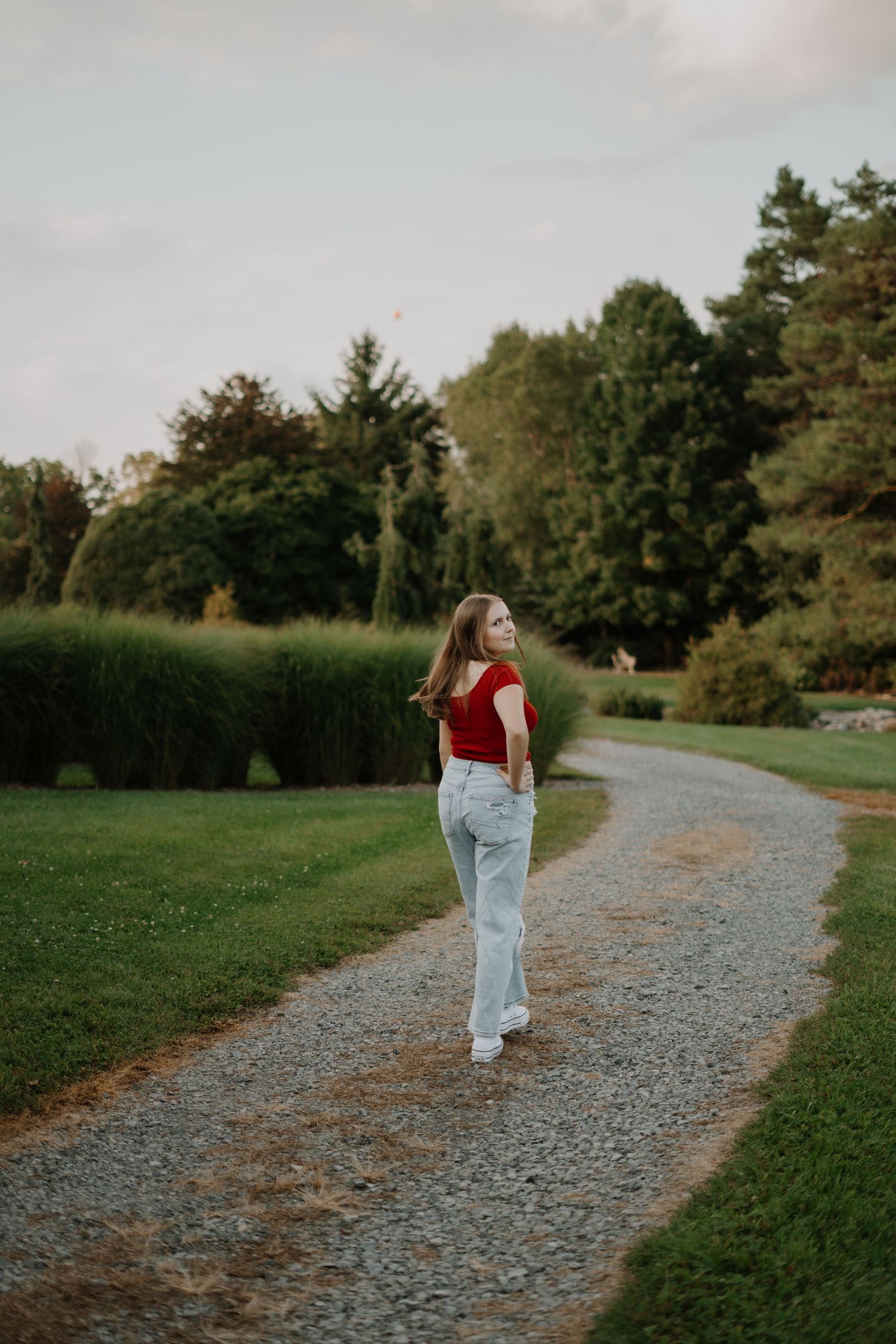 Senior portrait of person in red top and jeans walking along gravel path through green park setting.