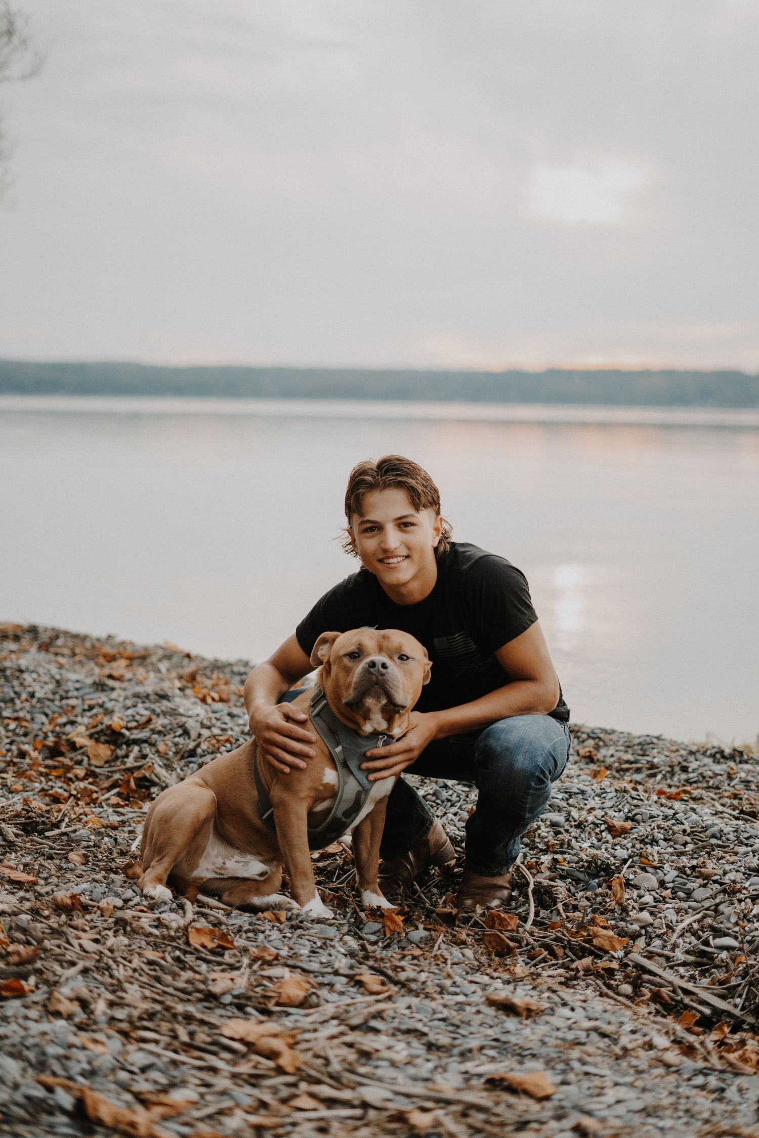 A senior portrait of a boy kneeling beside a muscular brown dog on a rocky lakeshore at sunset.