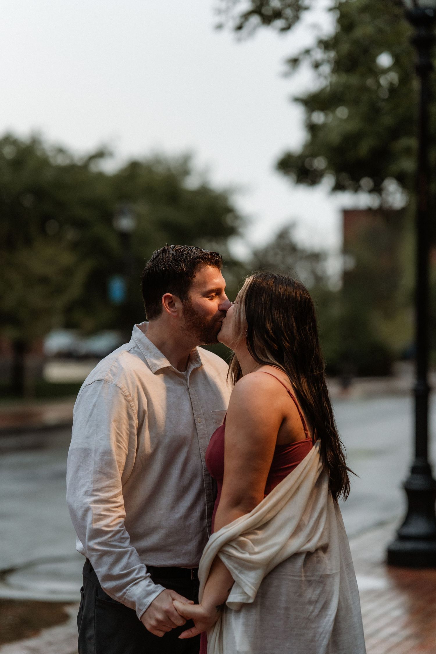 A couple shares a tender kiss while holding hands outdoors on a rainy day.