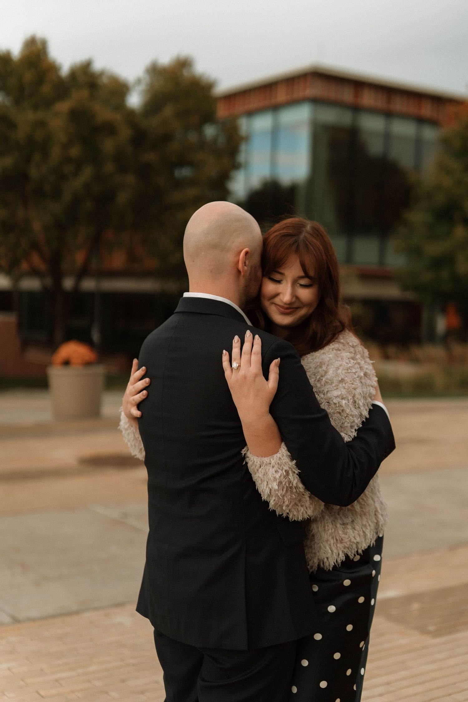 A couple shares a tender embrace outdoors, with the person in black hugging their partner wearing a polka dot dress.