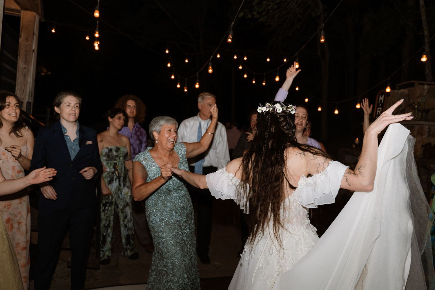 Bride in white gown dances joyfully with guests under string lights at an outdoor evening wedding reception.