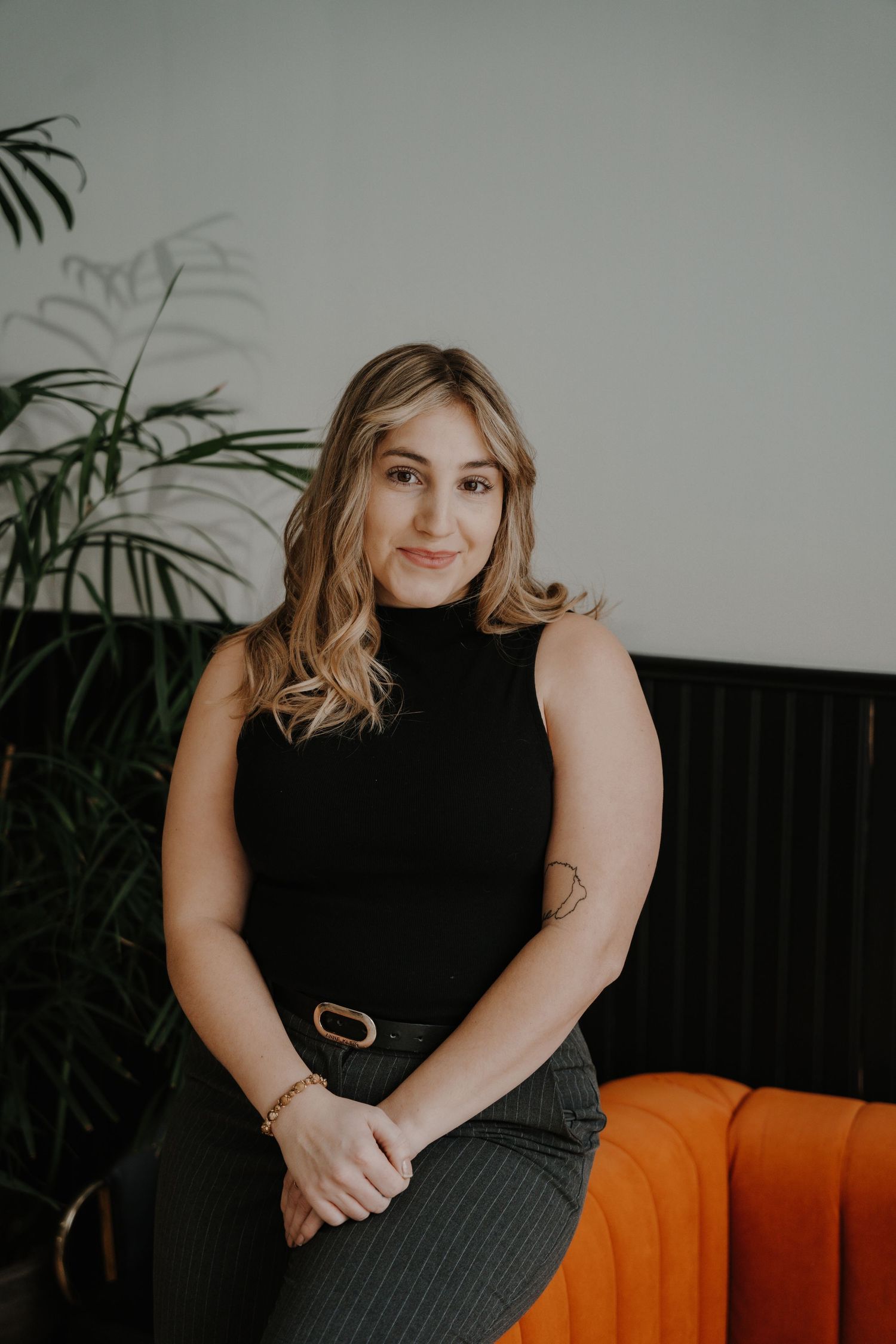 Headshot of a person in sleeveless black top sitting casually on an orange couch near indoor plants.