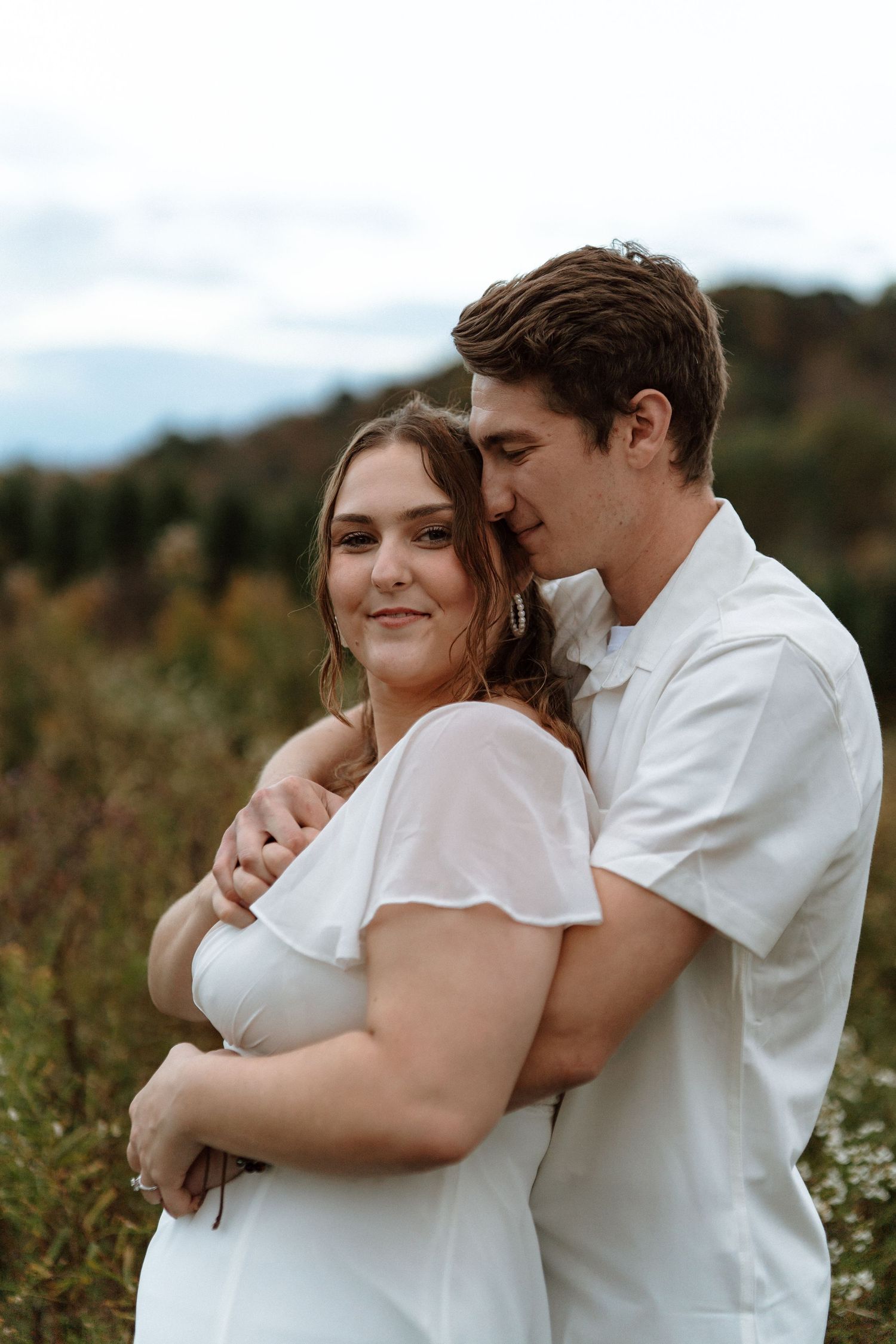 A romantic couple in white clothing embraces tenderly in a sunlit meadow during a photo session.