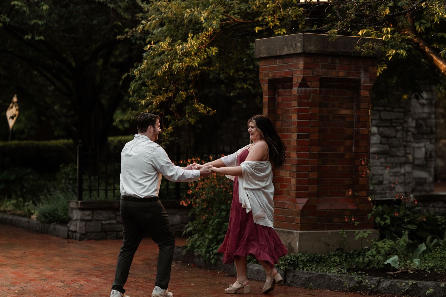 Couple dancing together on brick pathway in lush garden setting, woman in red dress, romantic outdoor engagement photo.