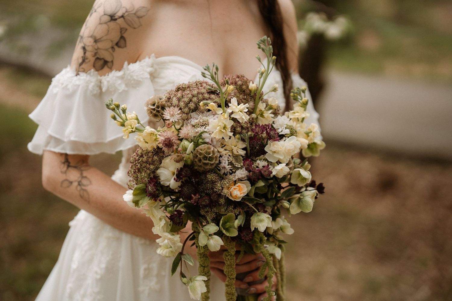 Tattooed bride in off-shoulder white gown holding lush cascading wedding bouquet with mauve, cream, and burgundy blooms.