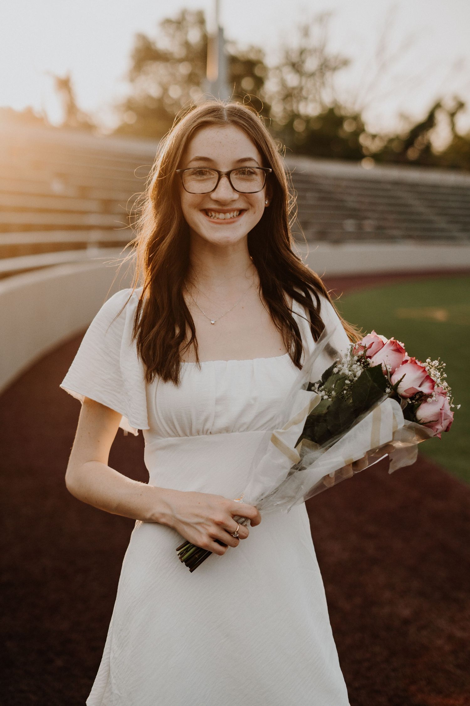 High school graduate in white dress holding bouquet of pink roses at outdoor stadium setting.
