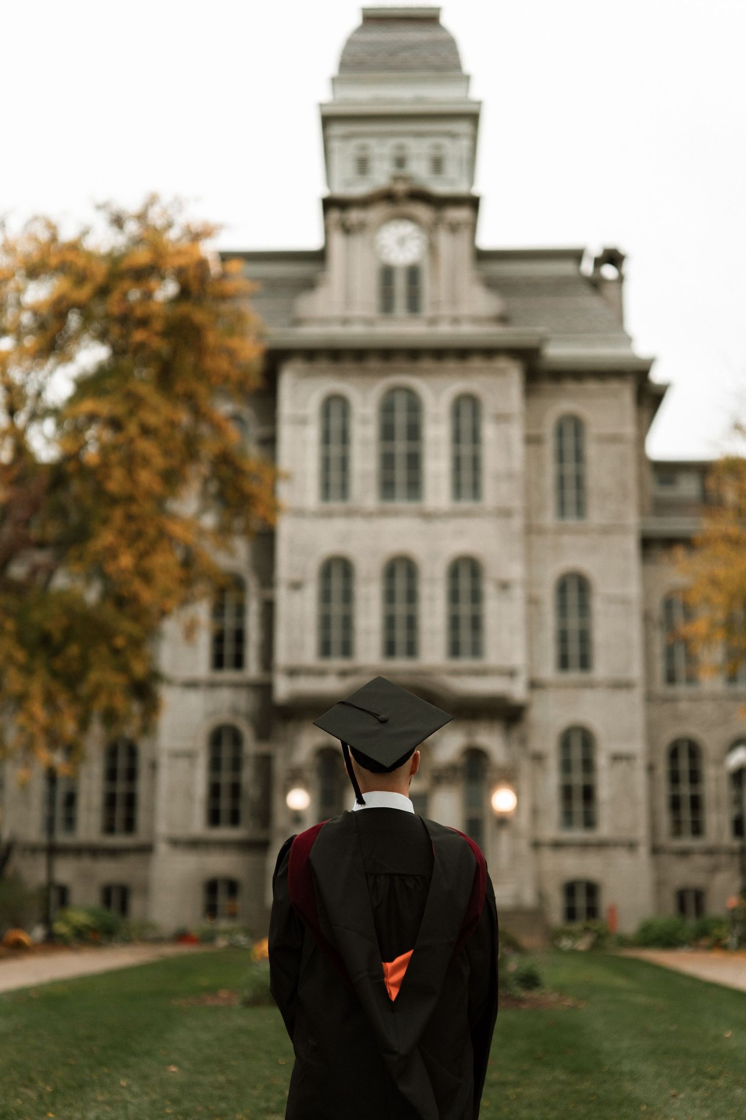 A historic stone university building with tall windows and towers stands behind a manicured lawn on an autumn day.