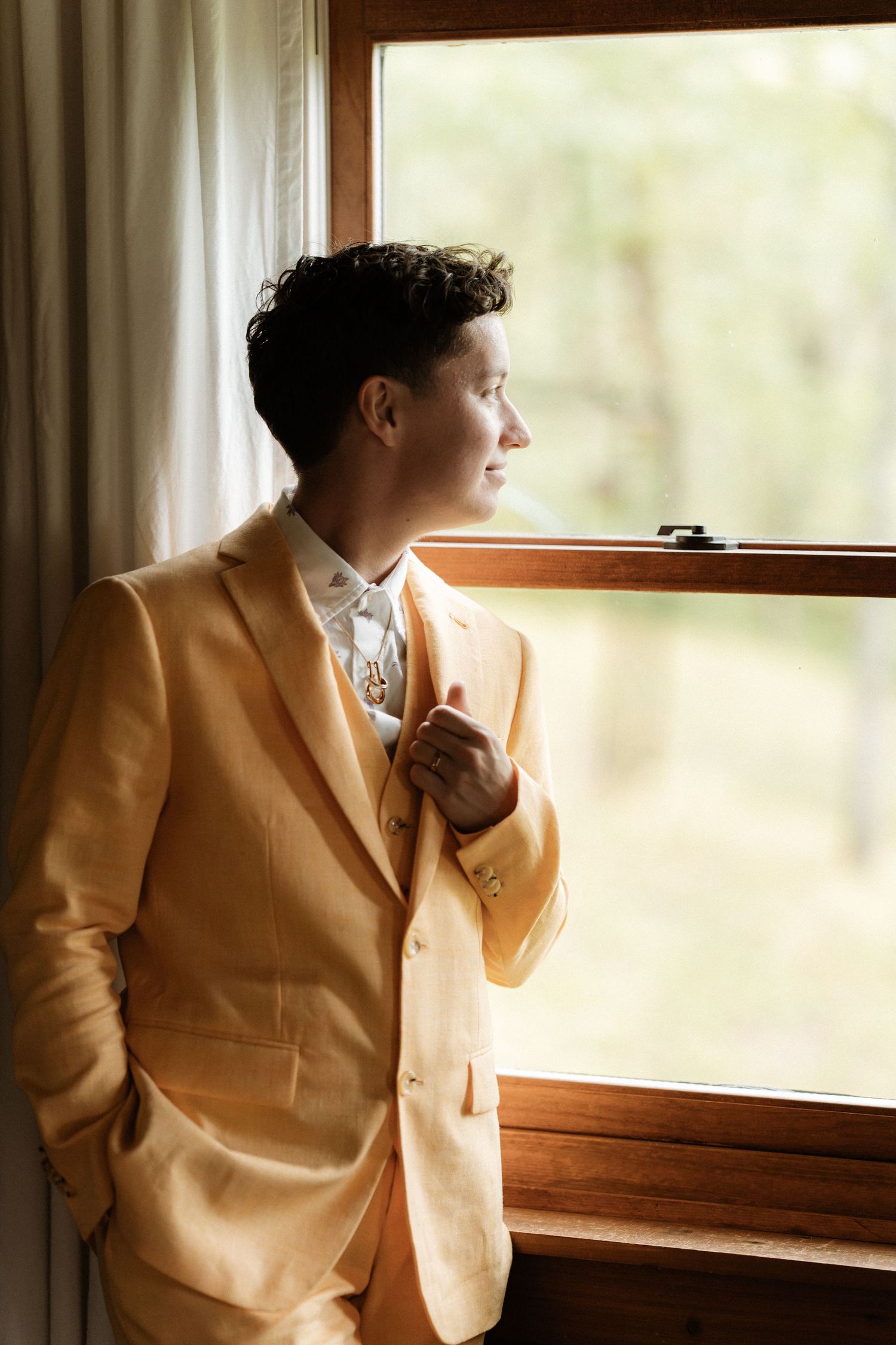 Groom in yellow suit gazing out wooden window, holding lapel, with soft natural light and green trees in background.