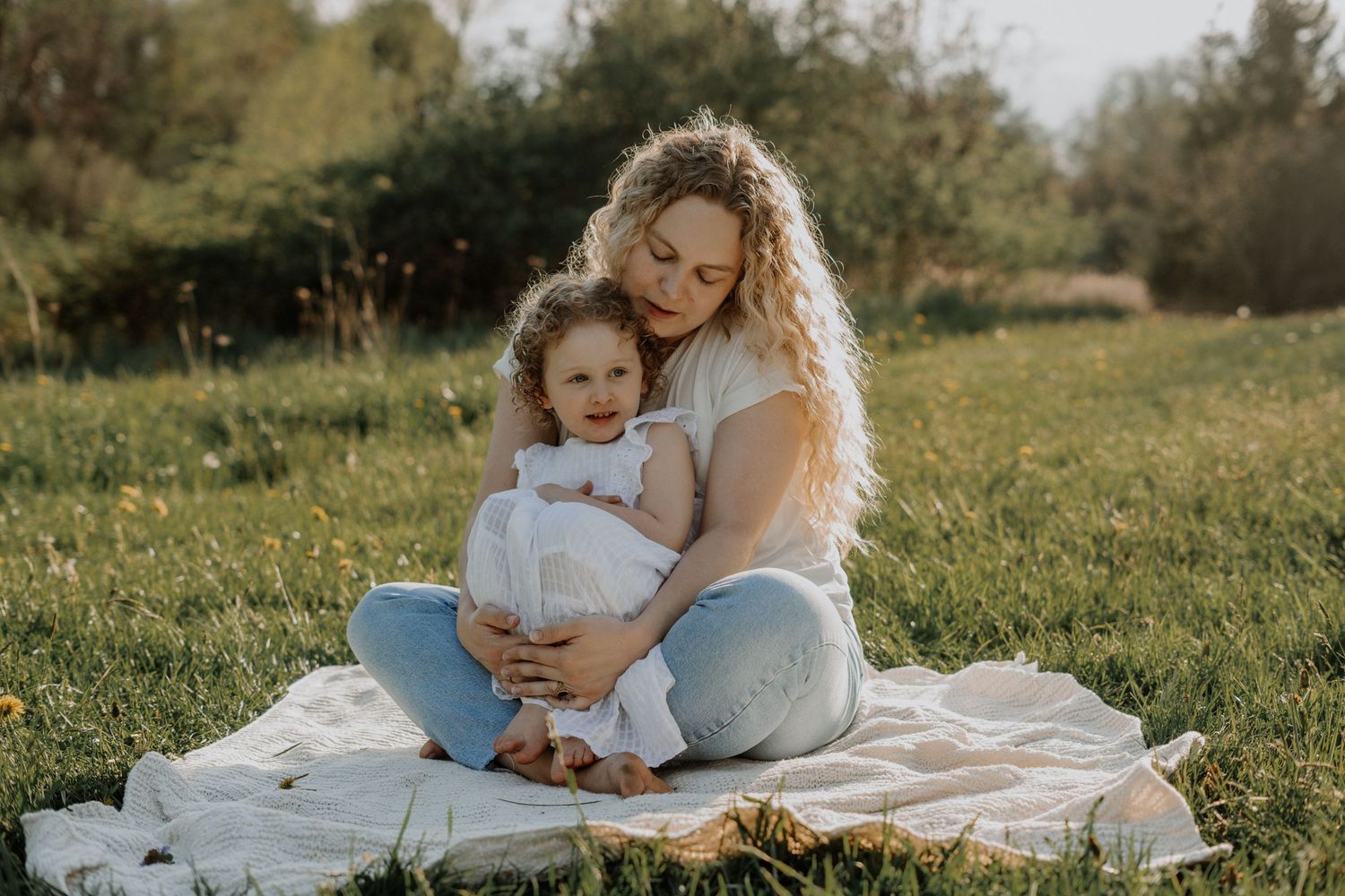 Parent and child share intimate cuddle while sitting on blanket in sunny field.
