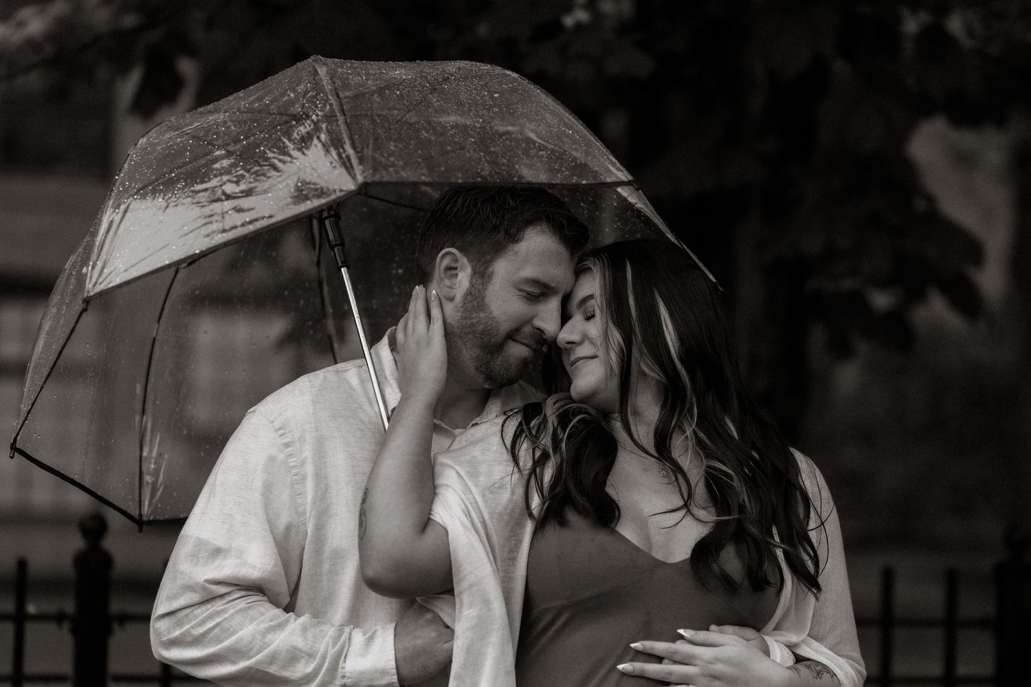 Black and white photo of a couple sharing a tender moment under a clear umbrella in the rain.
