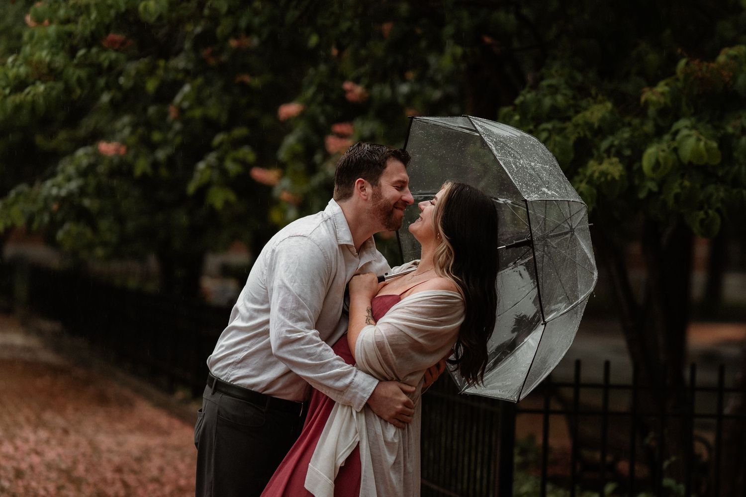 Couple sharing a romantic moment under a clear umbrella in the rain, surrounded by lush green trees.