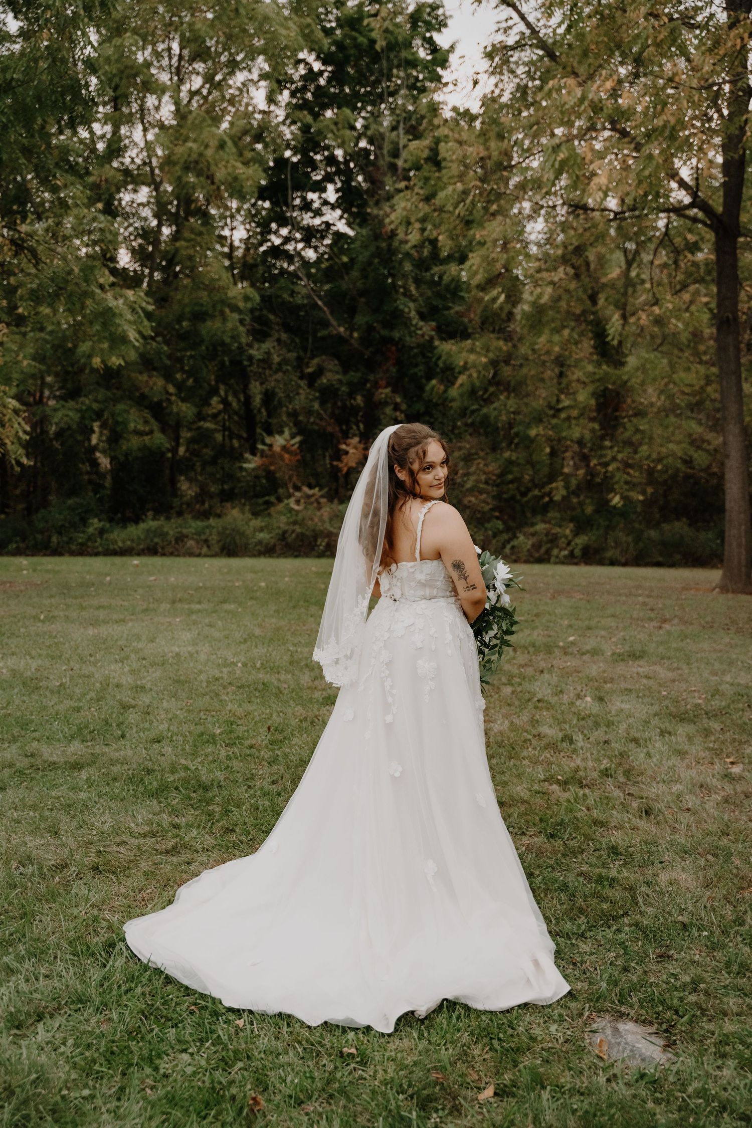 Bride in flowing white dress poses in natural outdoor setting with trailing veil.