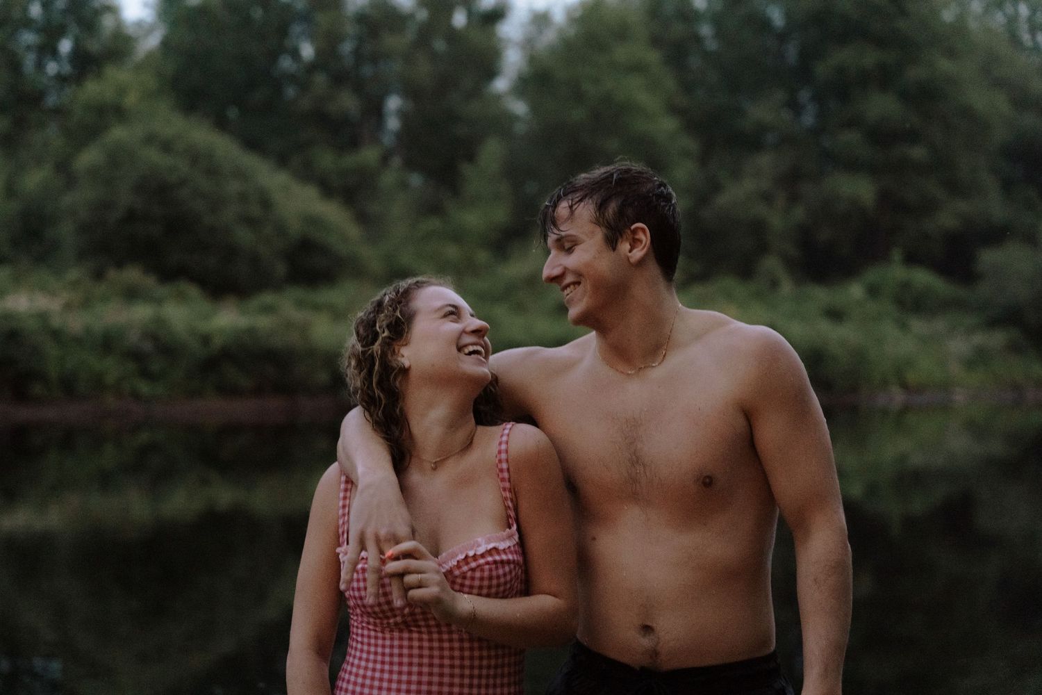 Couple in casual summer attire sharing intimate moments by a serene lake surrounded by lush greenery.