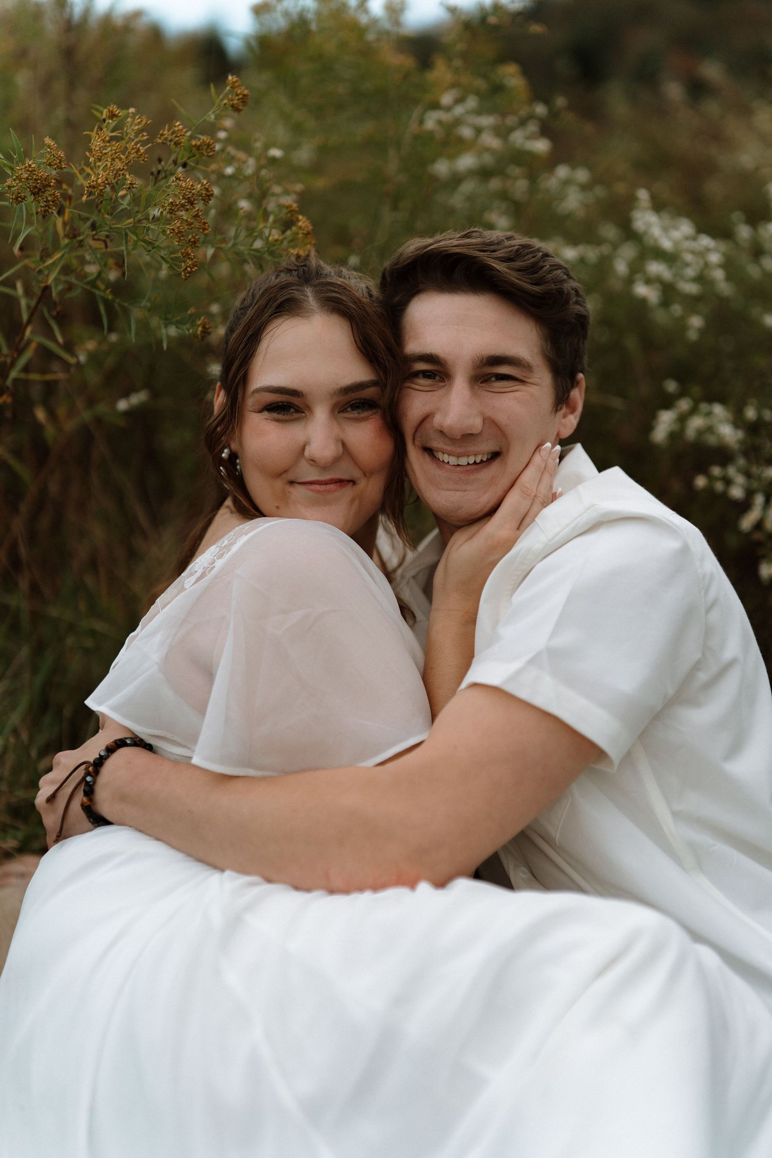 A romantic couple embraces outdoors in white clothing against a natural backdrop of greenery and flowers.