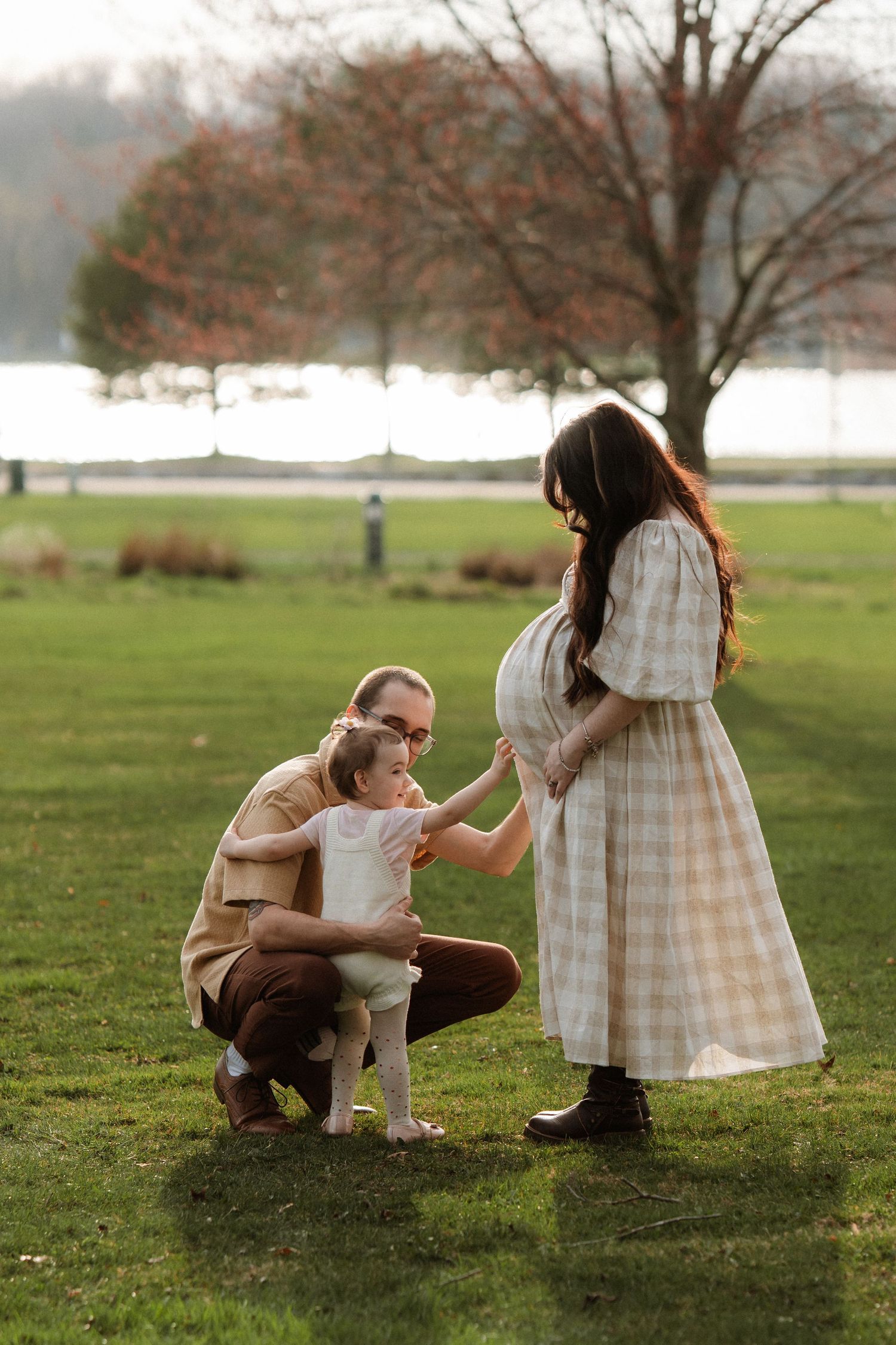 Pregnant woman in plaid dress with husband and toddler enjoying a family moment outdoors in a green park.