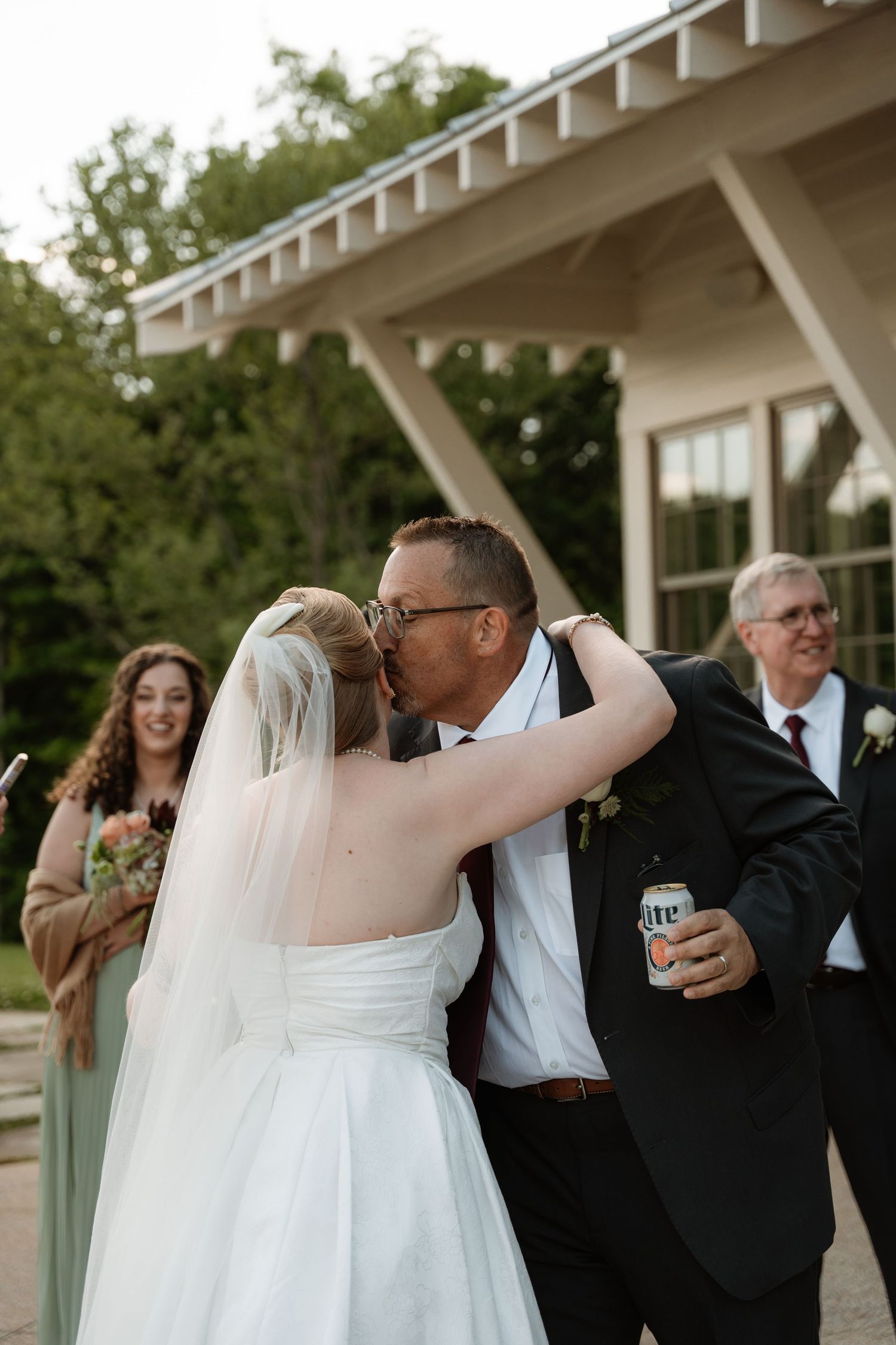 Bride and groom share first kiss outdoors at wedding ceremony, guests smiling in background near modern building.