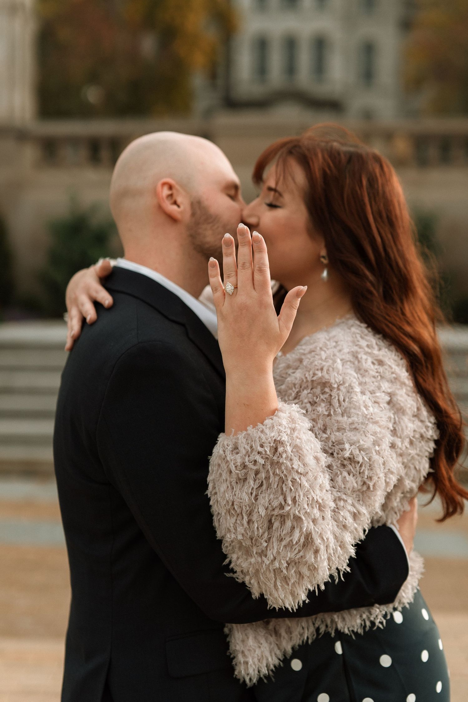 Couple sharing a romantic kiss outdoors, woman showing engagement ring in fuzzy coat and polka dot dress.