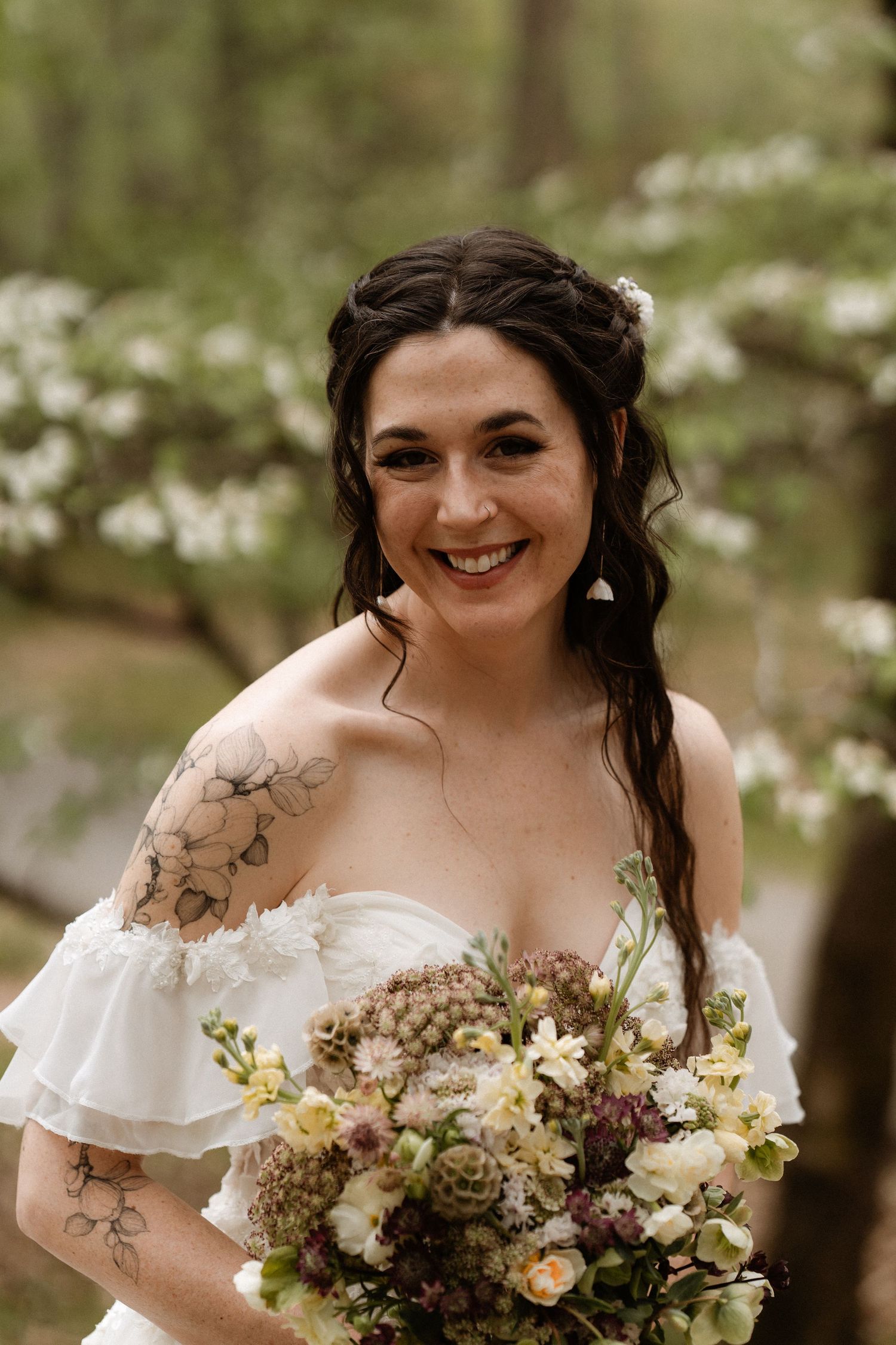 Smiling bride in white off-shoulder gown holds bouquet with succulents outdoors among blooming trees.