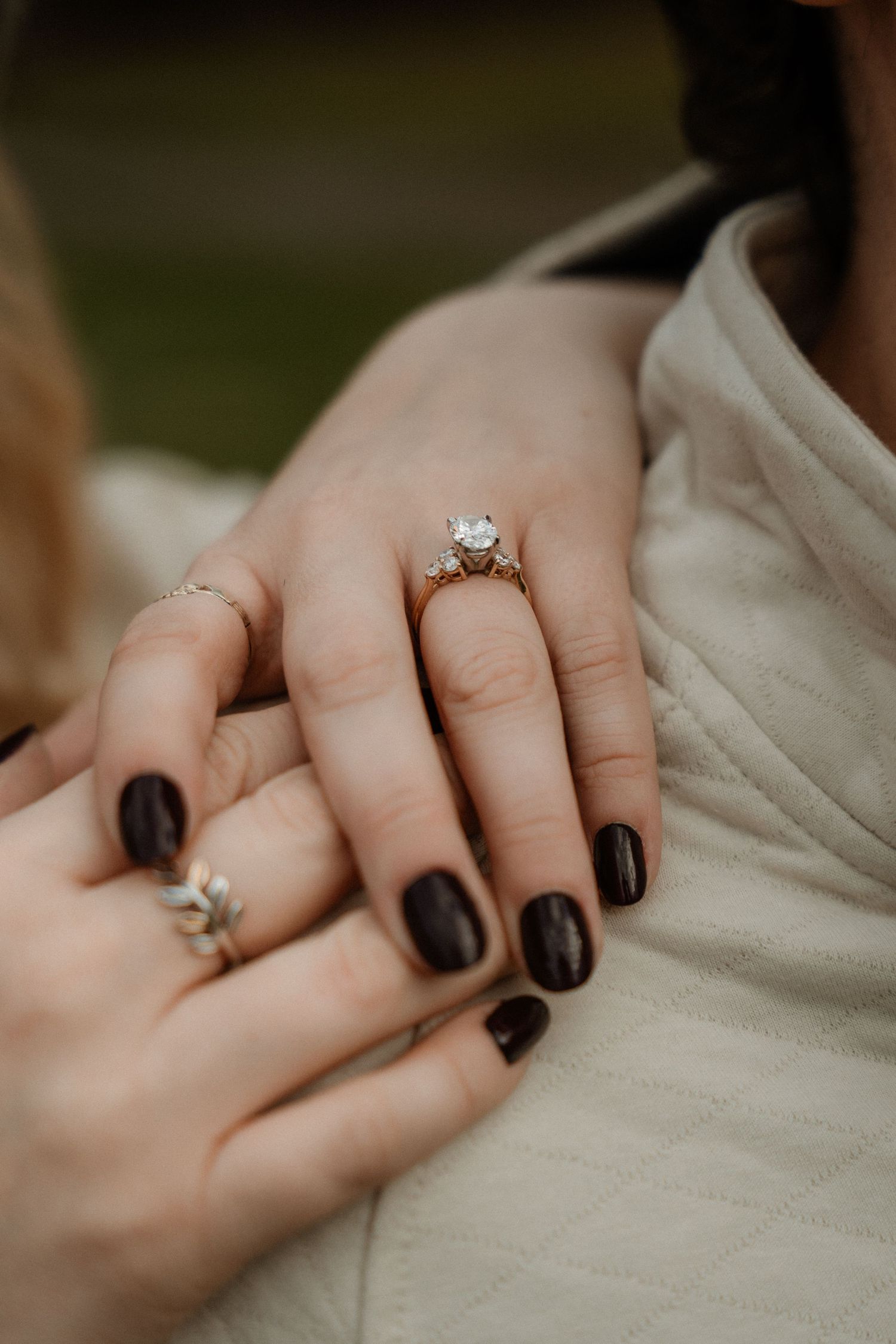 Close-up of two hands displaying elegant rings, including a diamond engagement ring and a floral band, with dark nail polish.