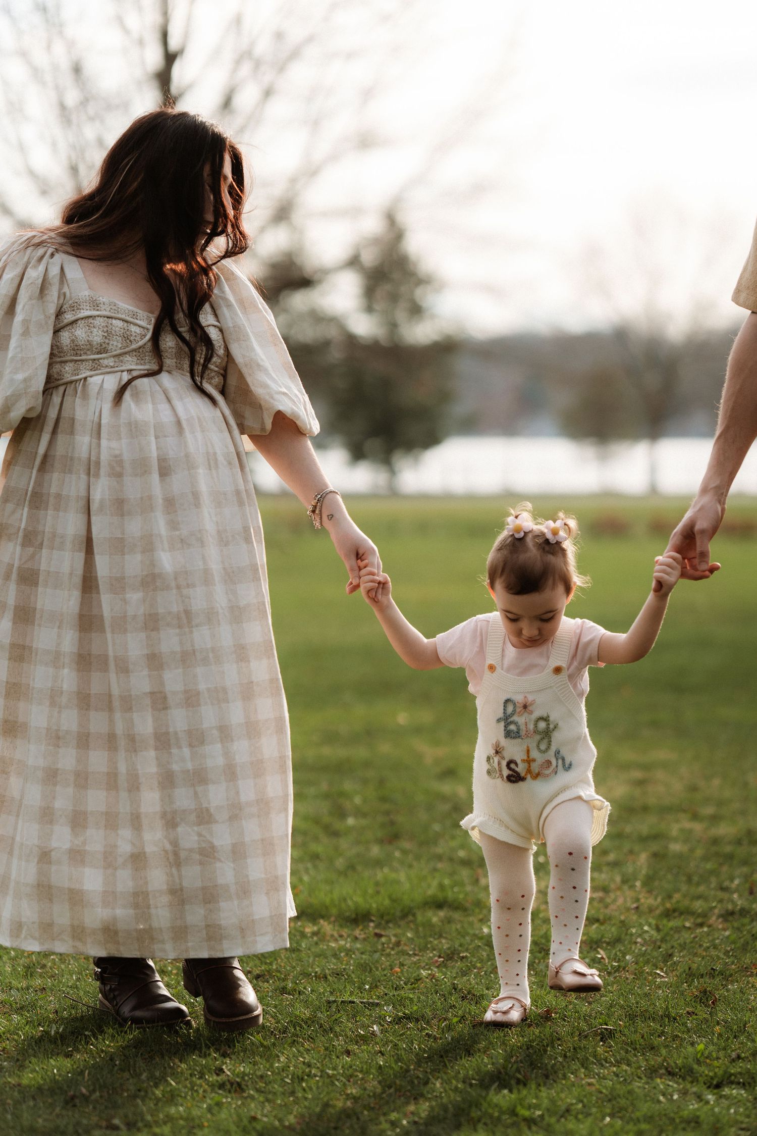 Pregnant mother and toddler girl in 'big sister' outfit holding hands with dad while walking on green grass.