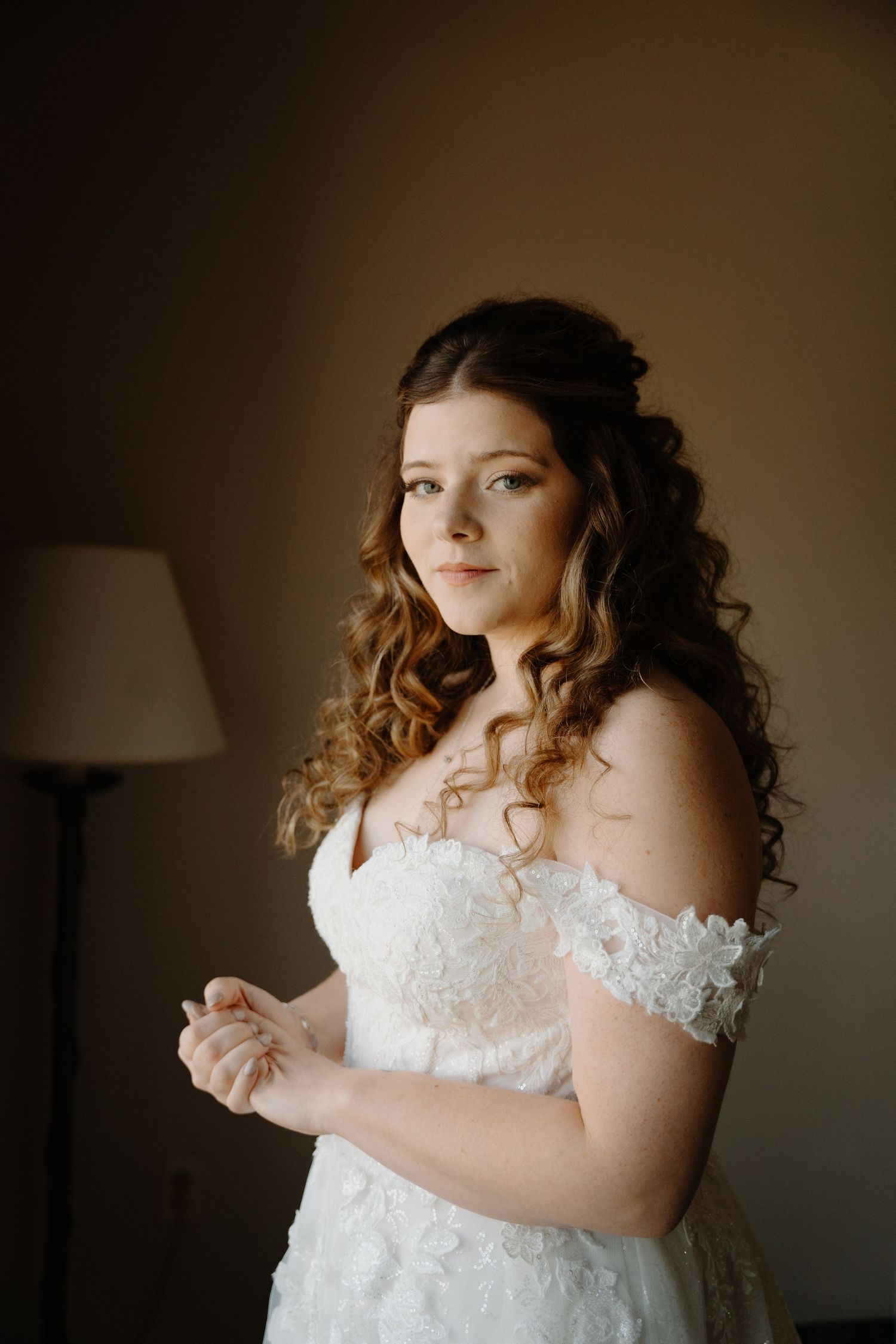 A bride in a lace off-shoulder wedding dress poses with curled hair in soft lighting.
