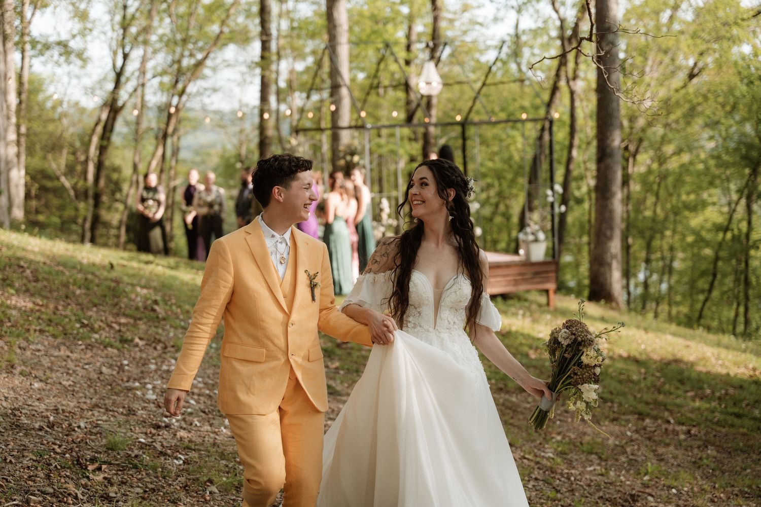 Bride and groom walk hand-in-hand through wooded outdoor wedding venue, groom in yellow suit, bride in white gown.