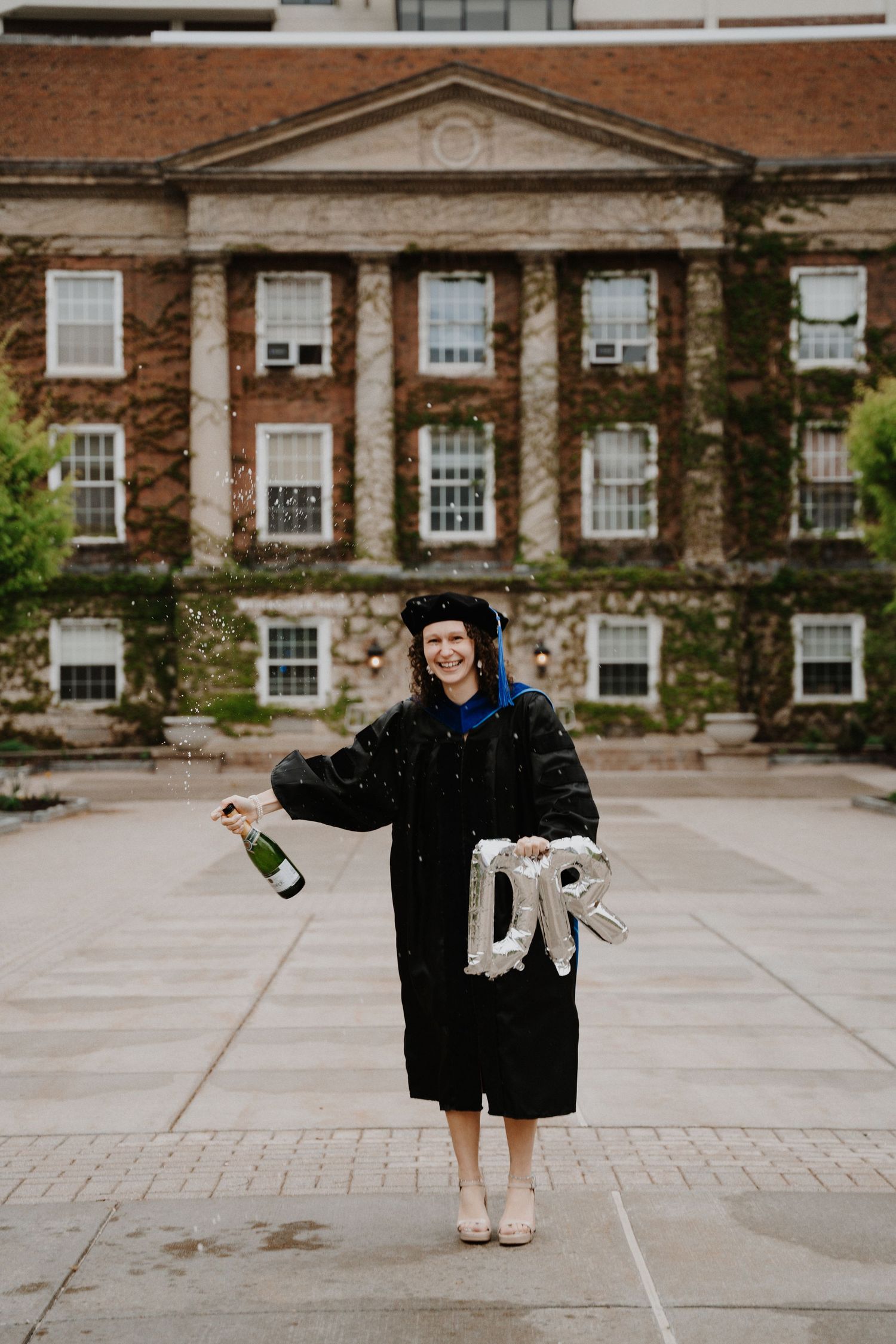 Graduate in traditional cap and gown celebrating with champagne in front of brick university building.