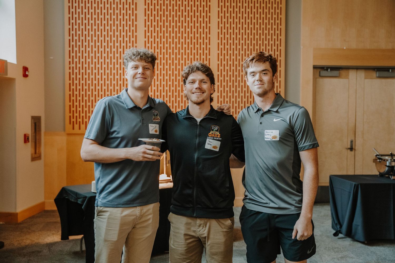 Three colleagues posing together at a professional event with a wood panel wall background.