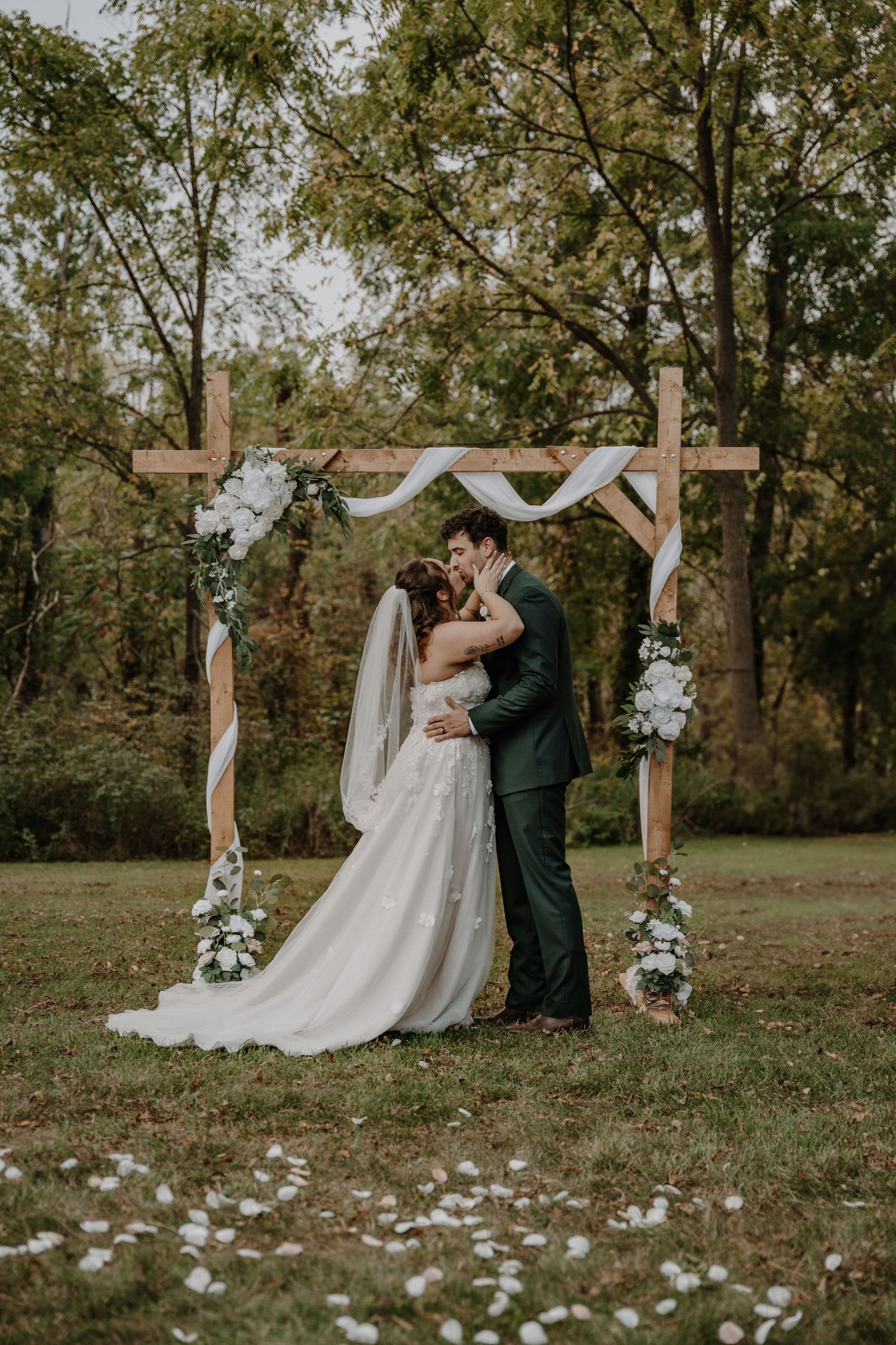Wedding ceremony takes place outdoors with wooden cross arch decorated in white flowers and greenery.