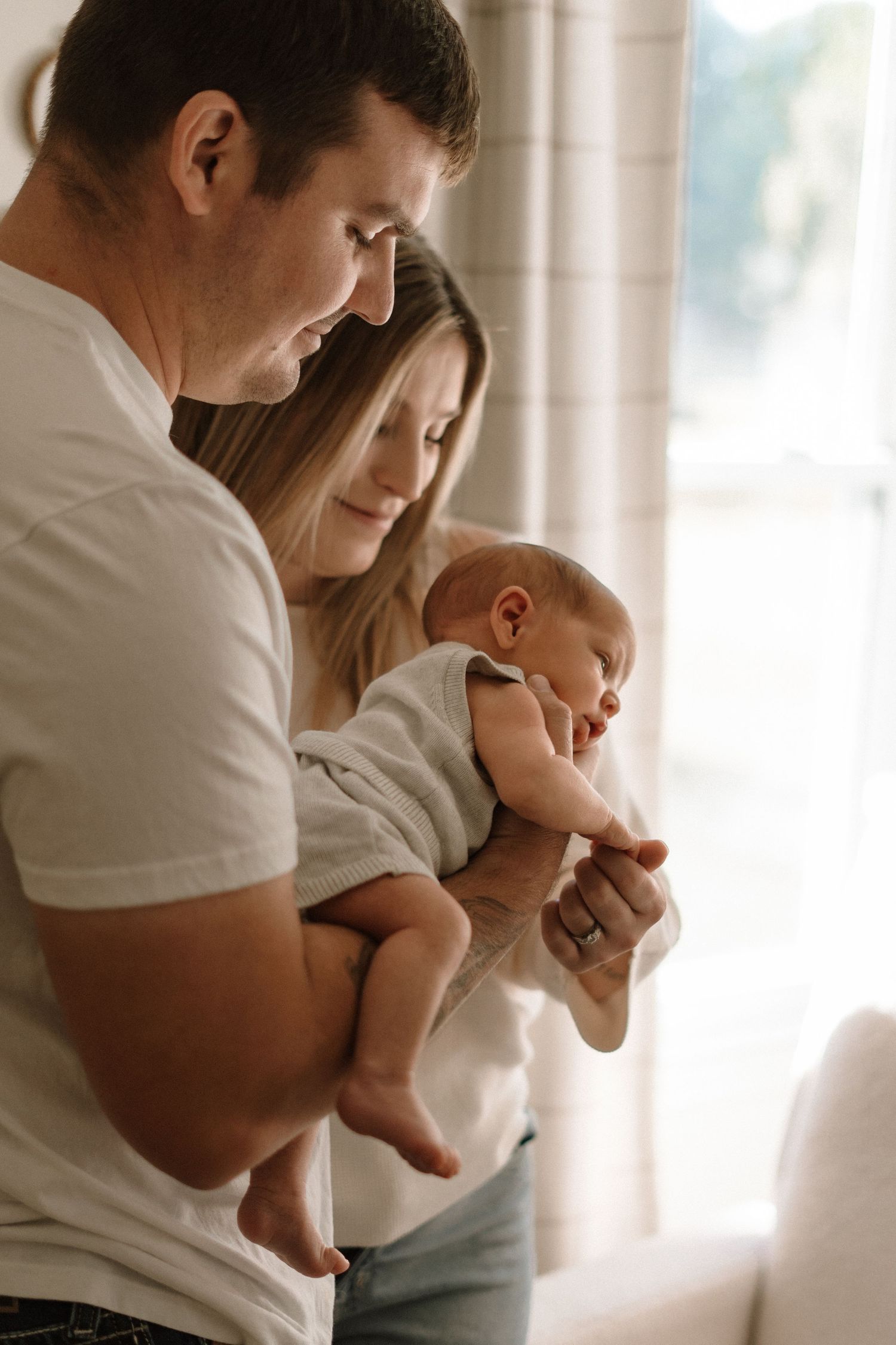 Parents in white clothing hold and gaze lovingly at their infant in a bright sunlit room.