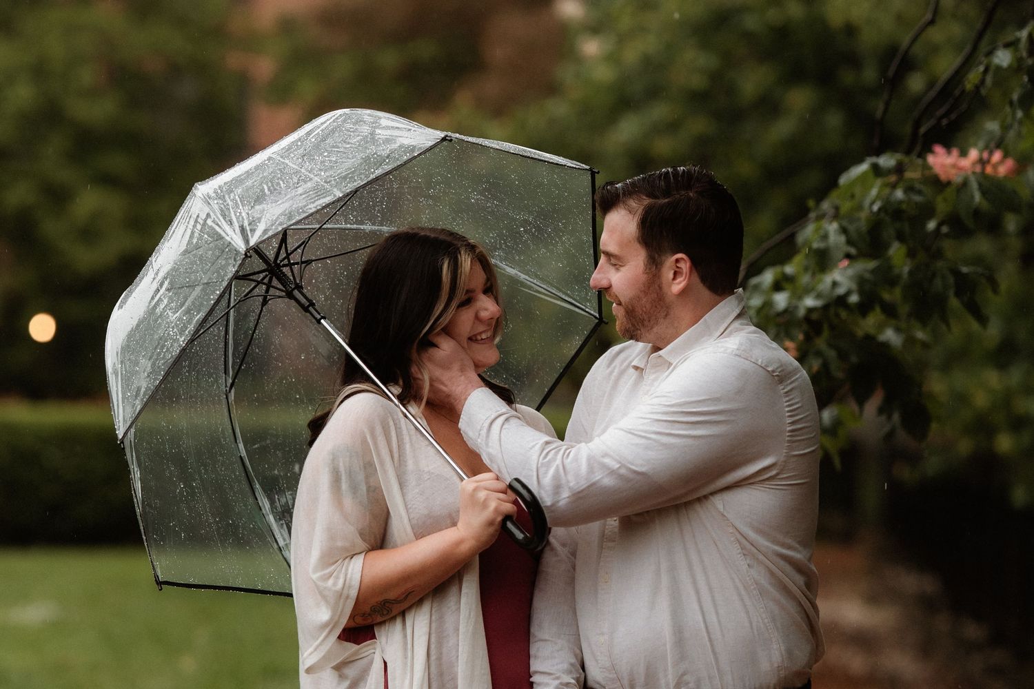 Couple sharing a clear umbrella in the rain, smiling at each other in a lush green garden setting.