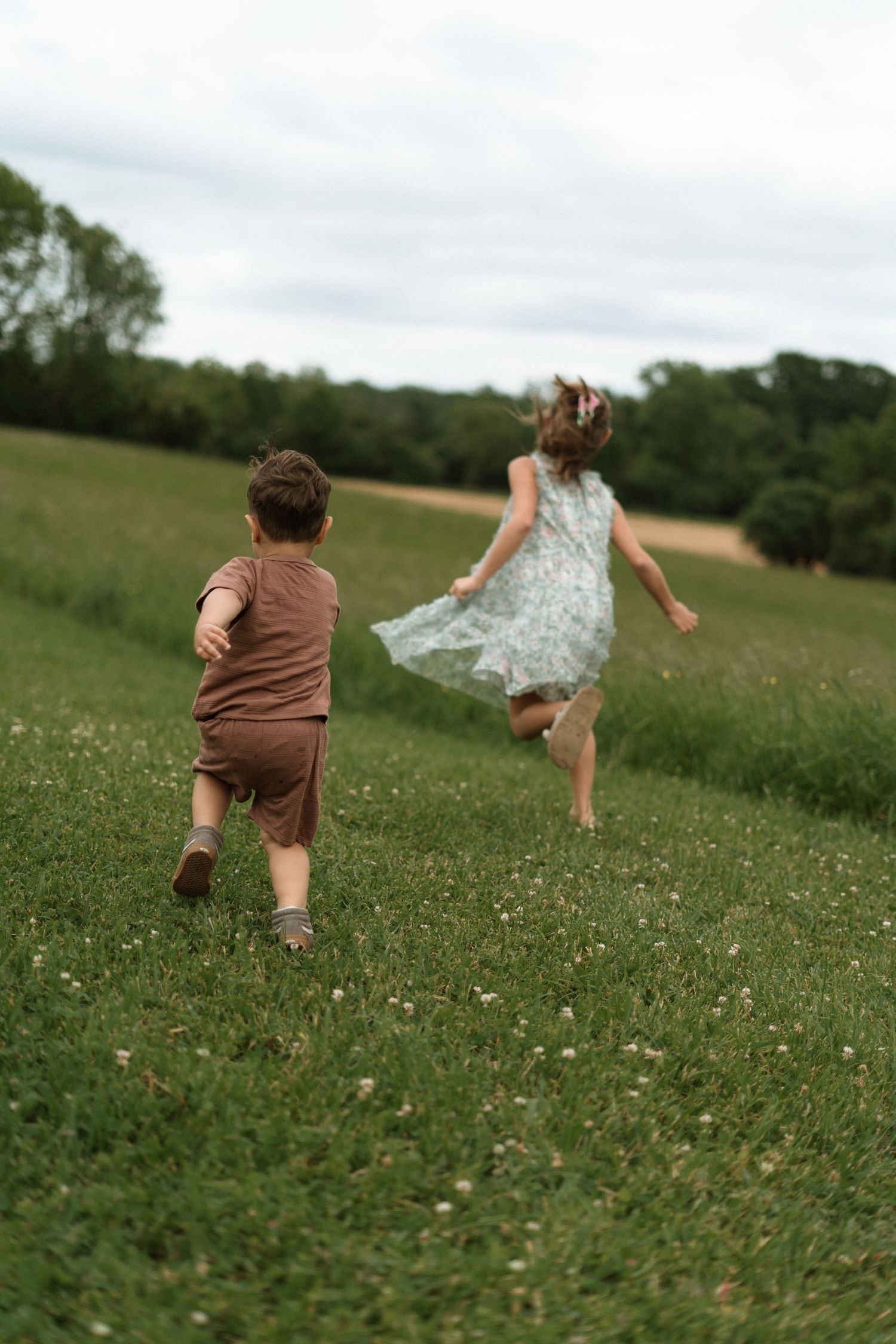 Two children run and play together across a lush green meadow dotted with wildflowers on a cloudy summer day.
