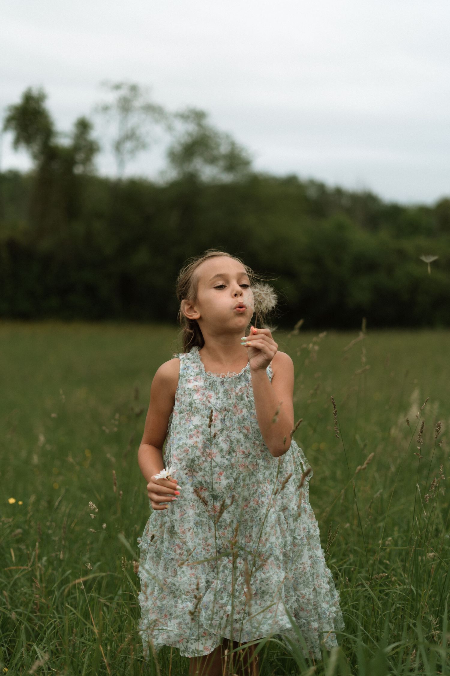 A young child in a floral dress blows on dandelion seeds in a summer meadow with trees in the background.