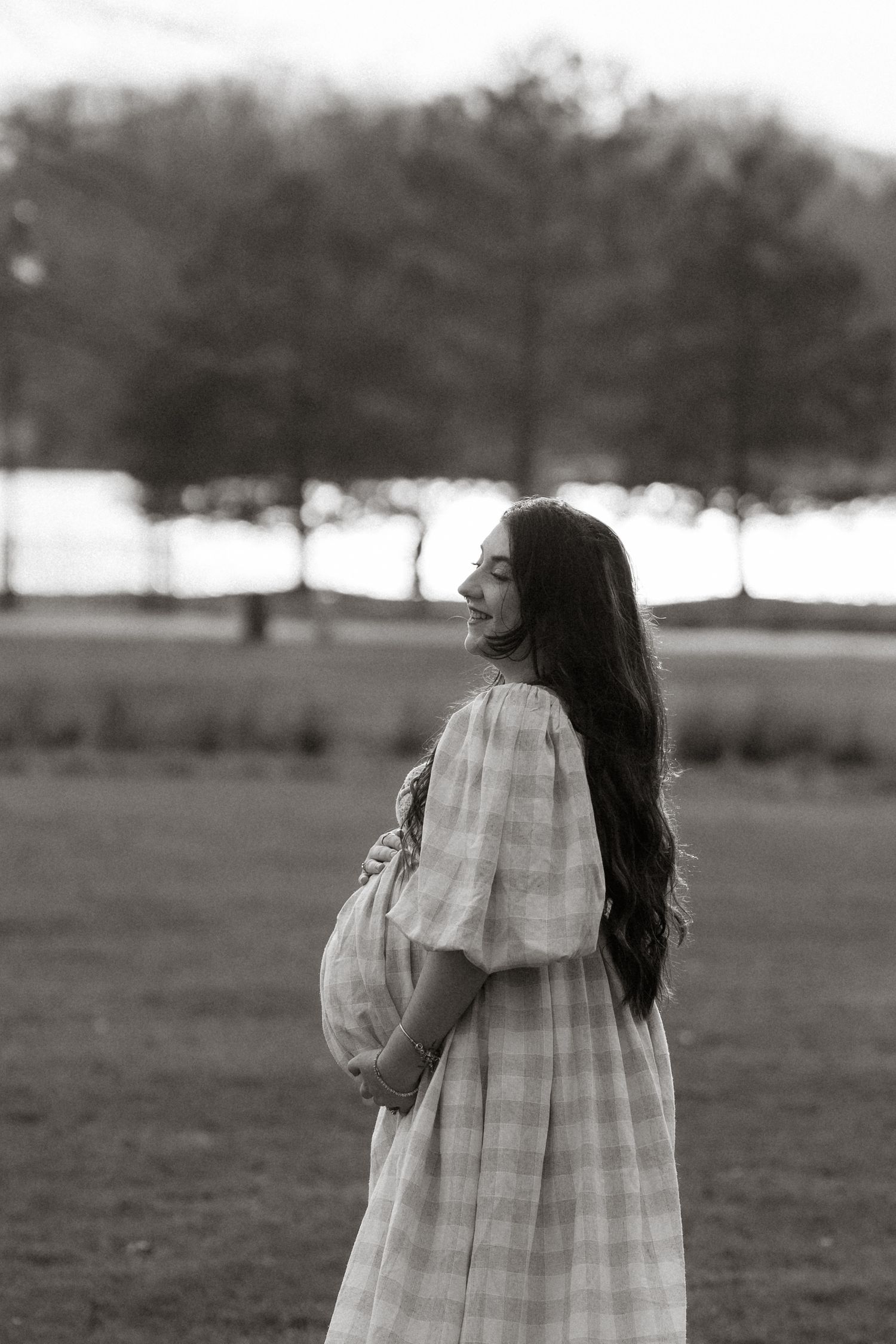 Black and white photo of a pregnant woman in a plaid dress holding her belly, standing in a park.