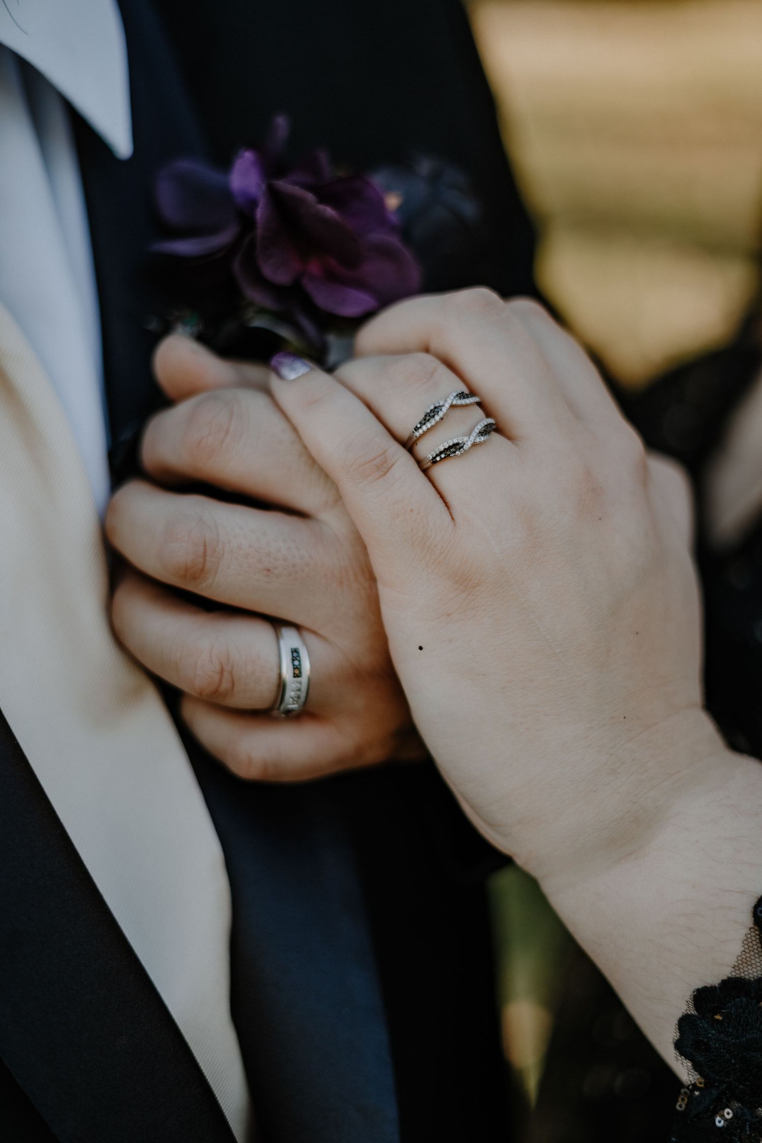 Close-up of newlyweds' hands with wedding rings and purple boutonniere against formal attire.