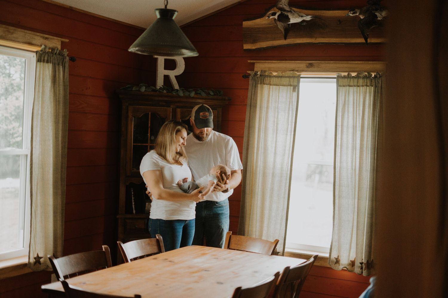 Interior of rustic dining room with wooden table and letter R wall decor. Couple with baby stand in front looking lovingly at newborn.