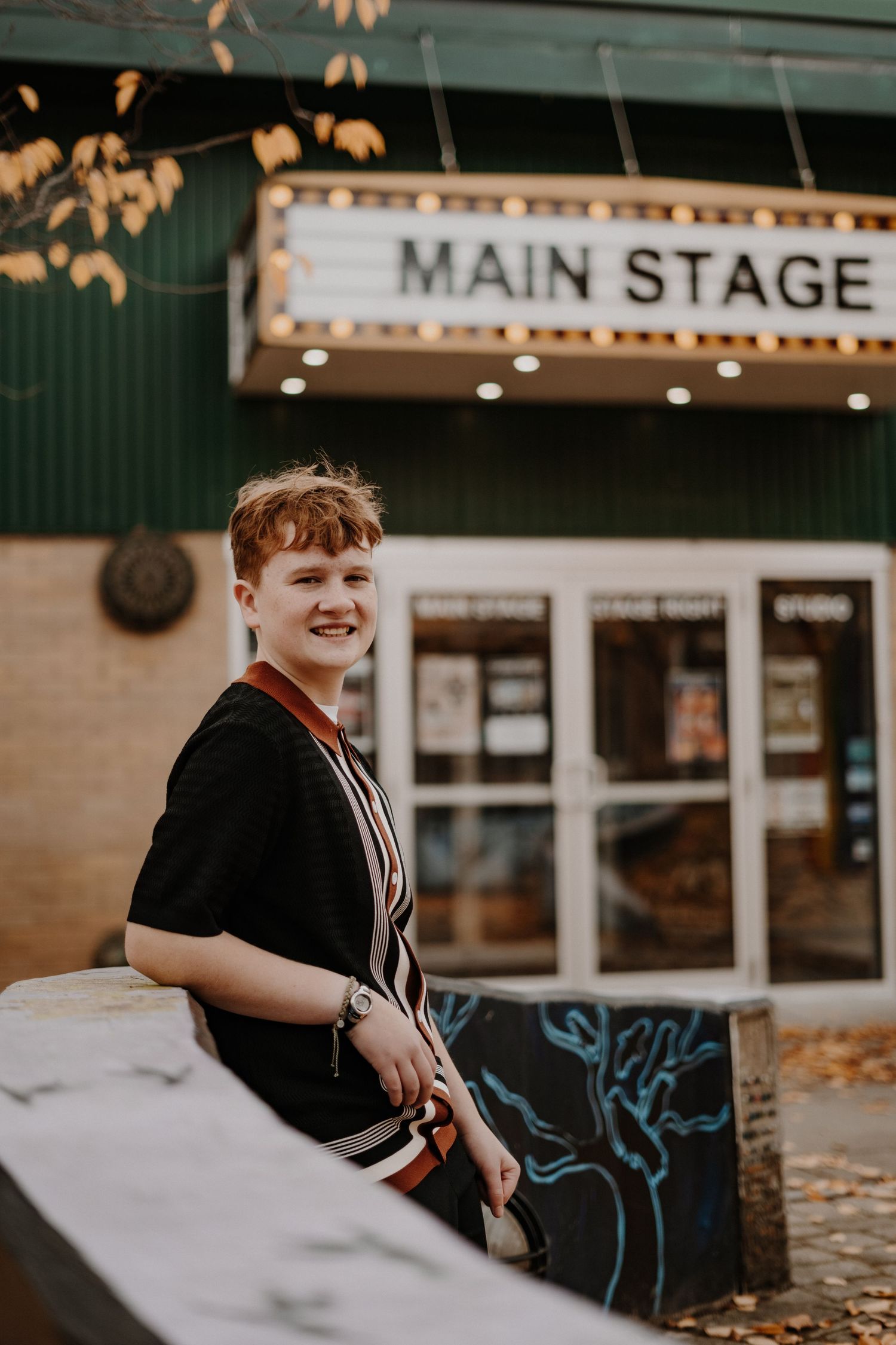 Senior portrait of person in casual attire sitting outside Main Stage theater entrance with autumn leaves.