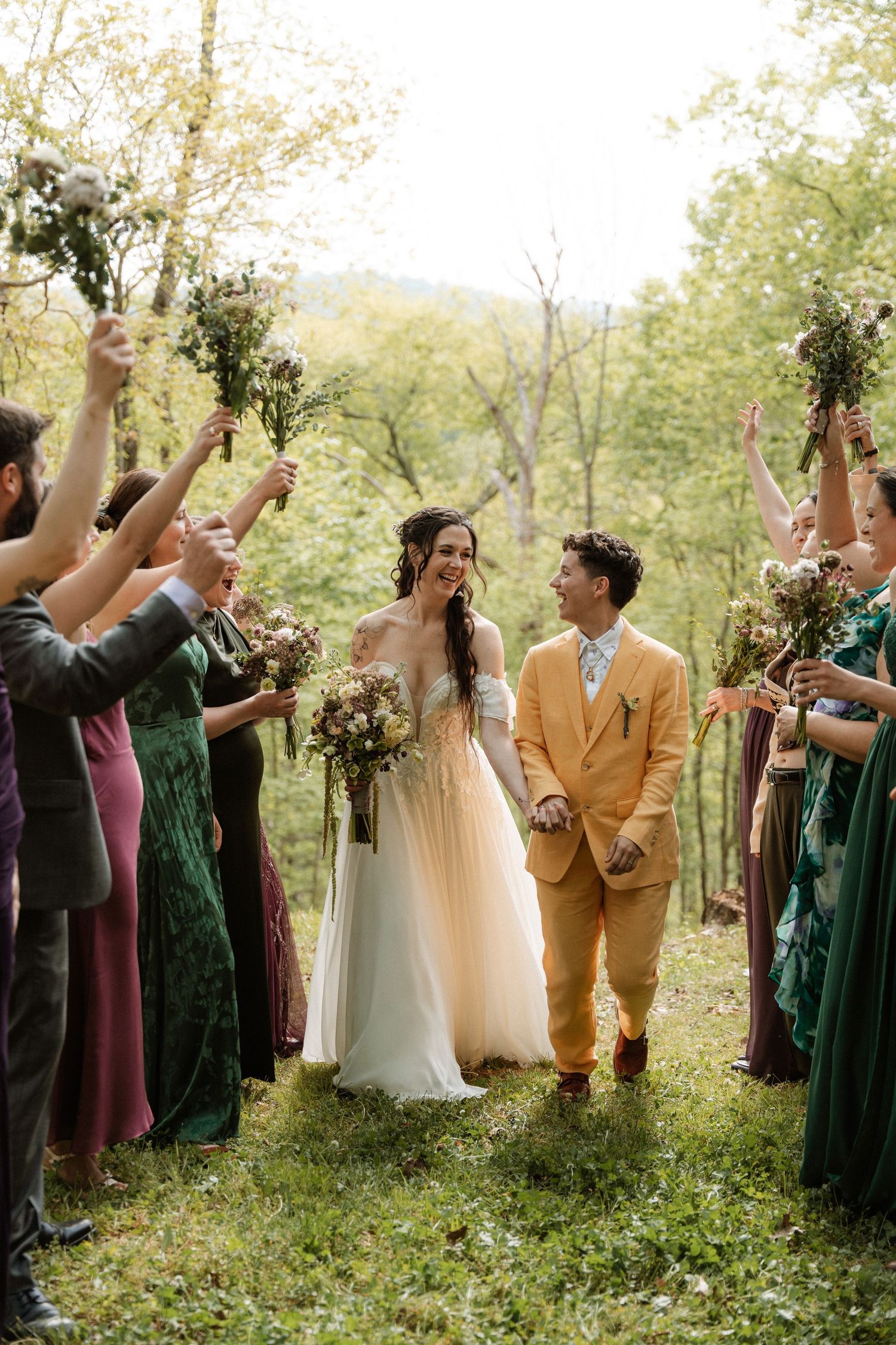 Couple walks through floral tunnel with wedding party in lush forest setting, bride in white gown, partner in gold suit.