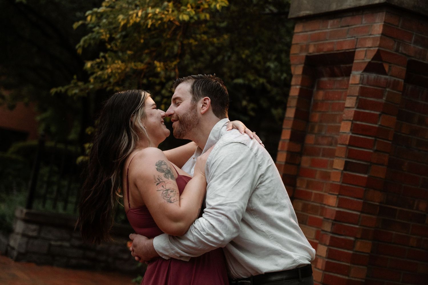 Couple sharing a romantic kiss outdoors near a brick wall, woman in burgundy dress with tattoo embracing man in white shirt.